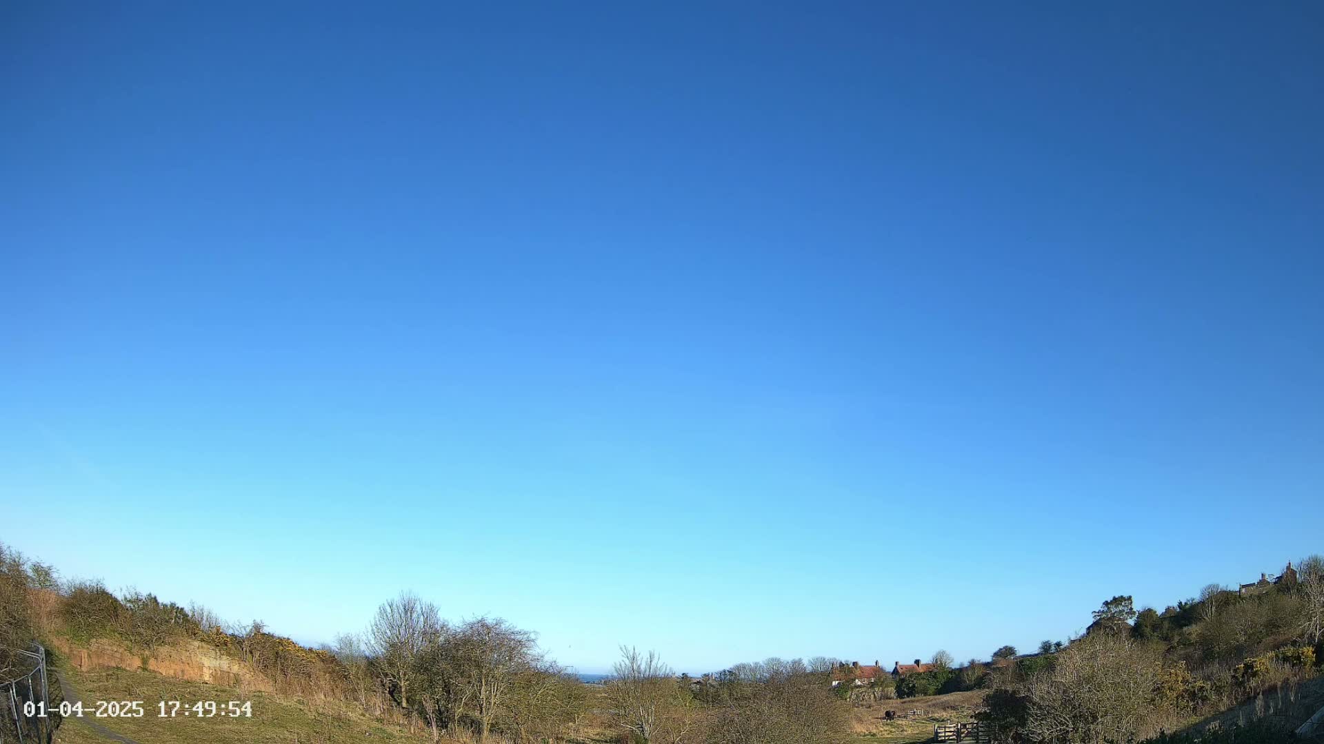 Under a clear blue sky, a pastoral landscape reveals a valley with sparse trees and a few distant buildings.