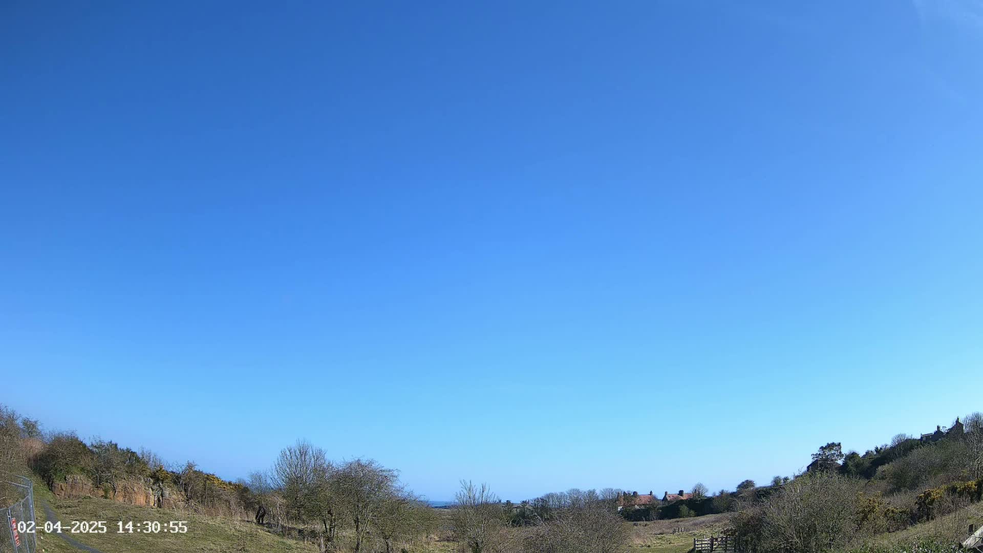 Under a clear blue sky, a hilly landscape with sparse trees and a few buildings in the distance is visible.