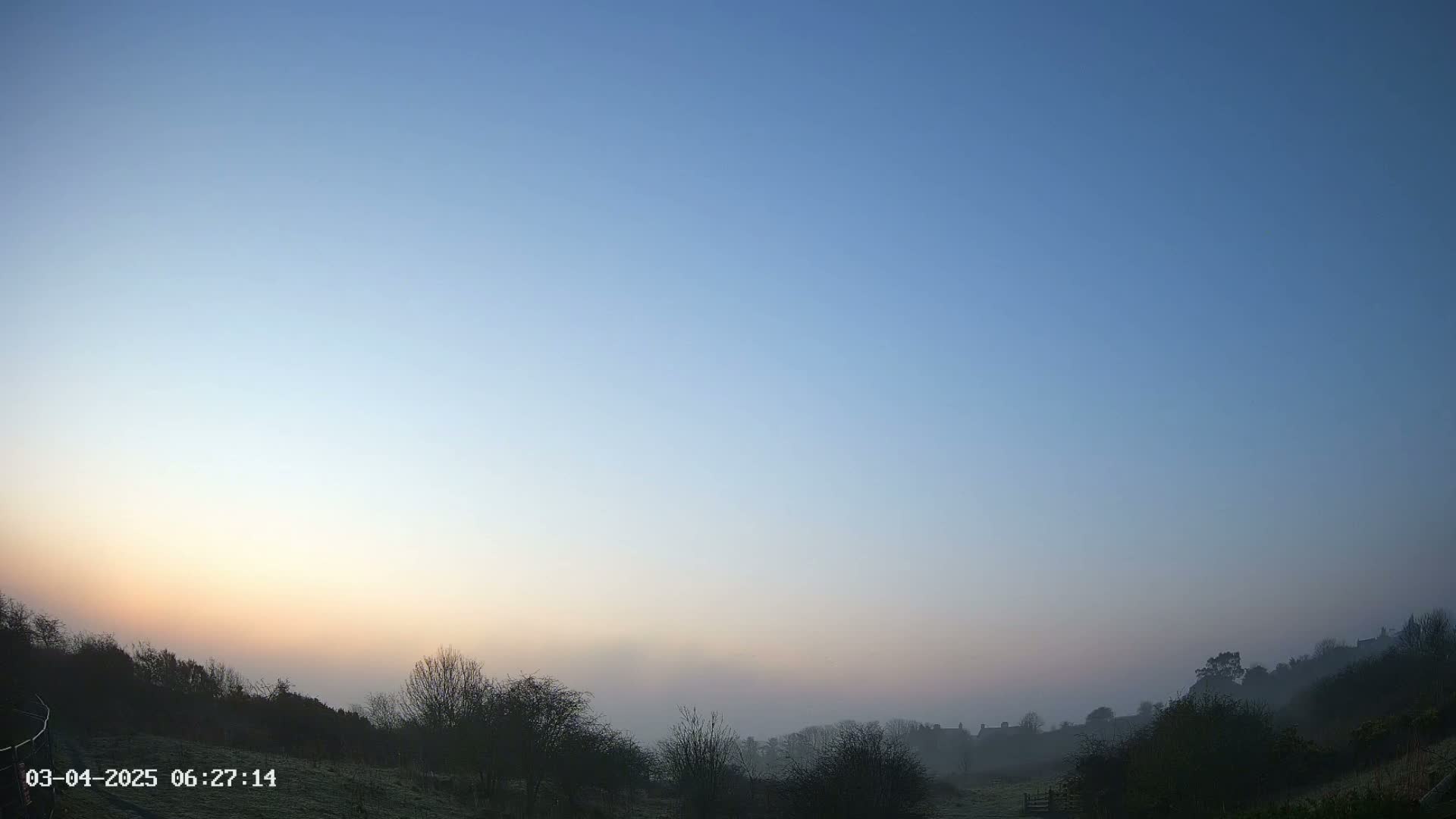 A misty sunrise illuminates a valley of trees and distant houses under a clear, pale blue sky.