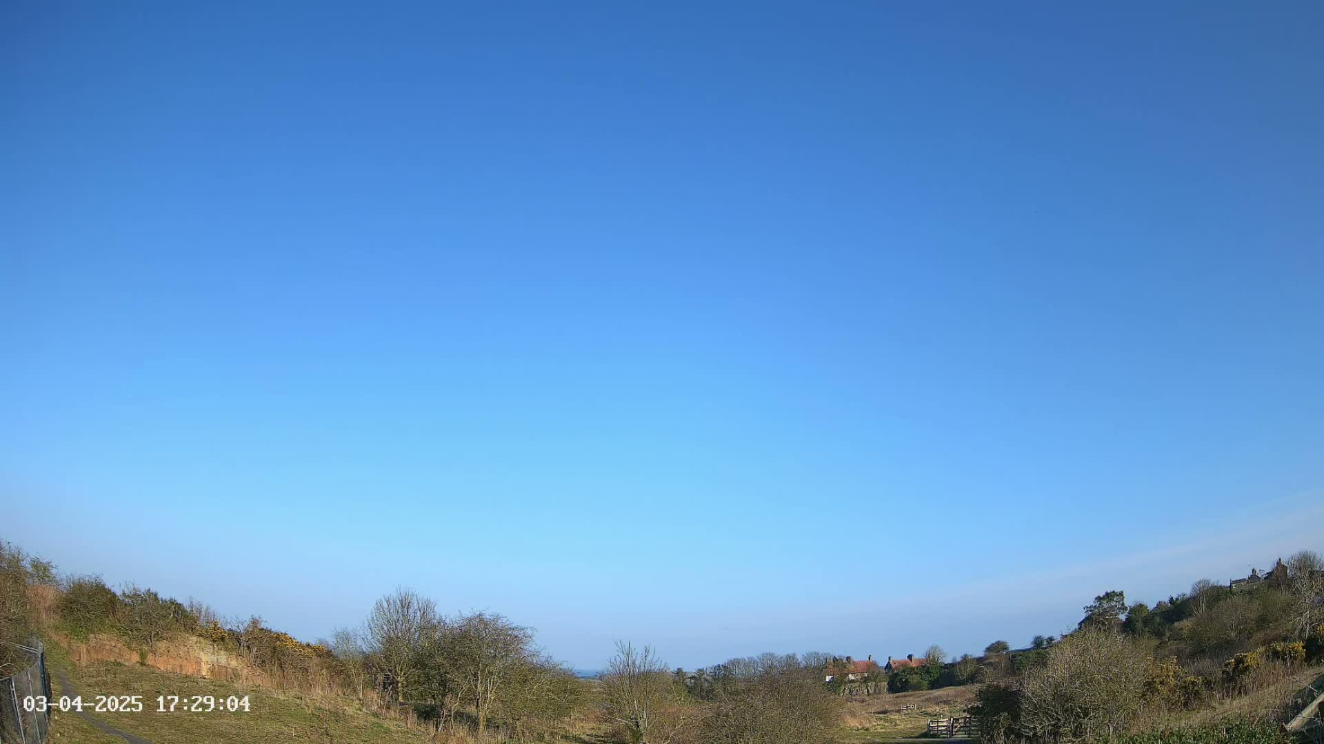 A clear blue sky overlooks a hilly landscape with sparse, leafless trees and a few buildings in the distance.