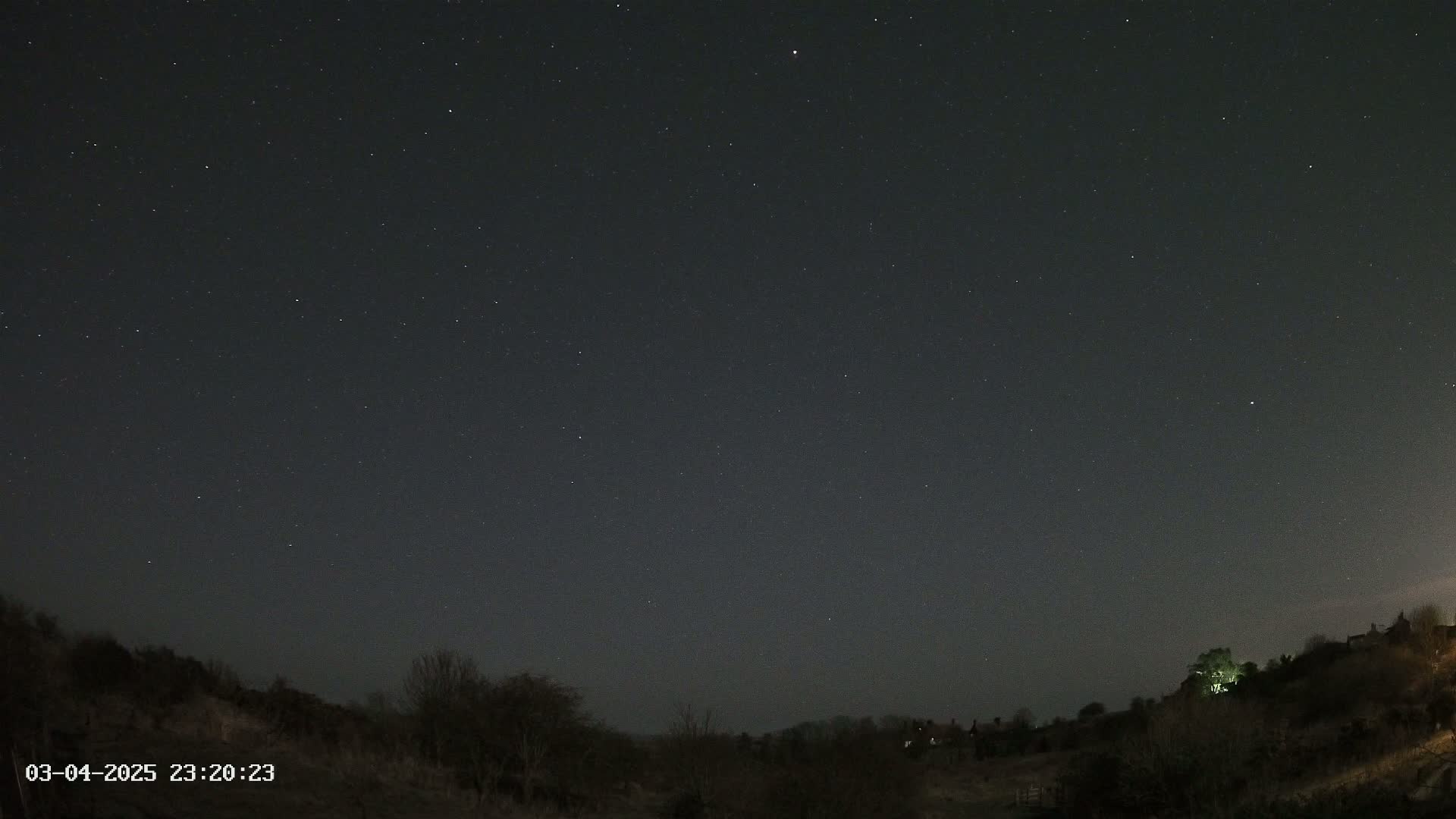 A night sky filled with stars is seen above a dark, hilly landscape with sparse trees and distant lights.