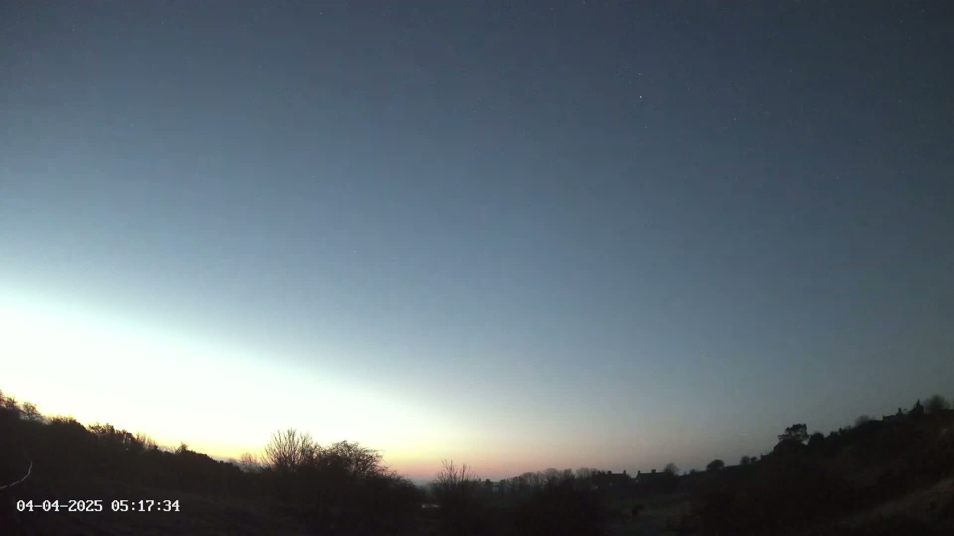 A clear, twilight sky with a faint pinkish-orange horizon line over a silhouetted landscape of trees and buildings.