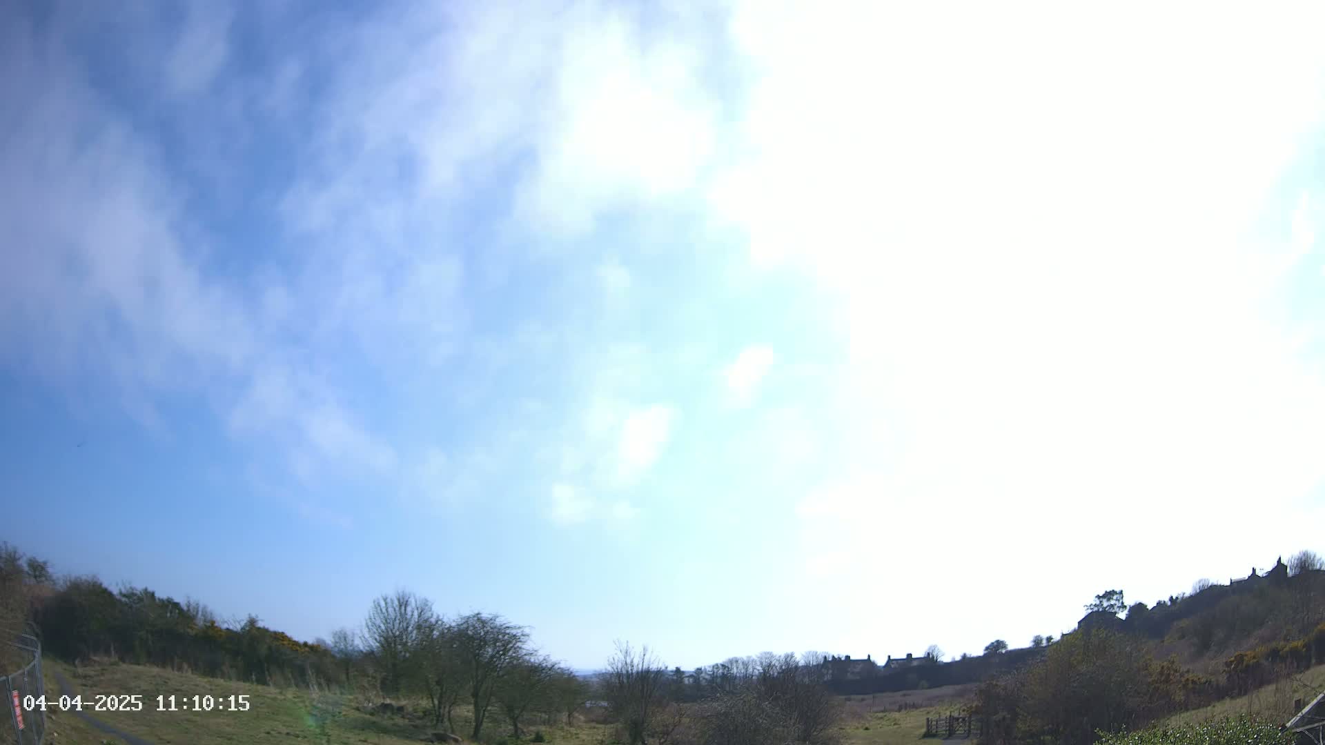 A mostly sunny day with scattered clouds displays a view of a grassy field with trees and distant houses on a hill.