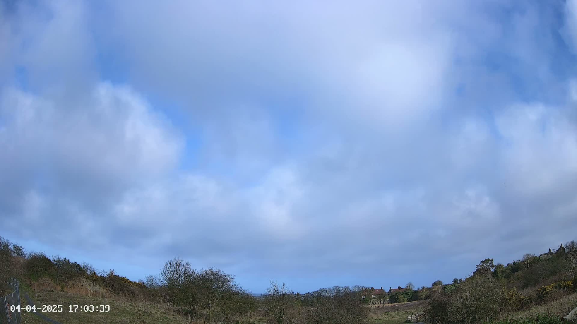 A mostly cloudy sky over a hilly landscape with sparsely wooded areas and a few houses in the distance.