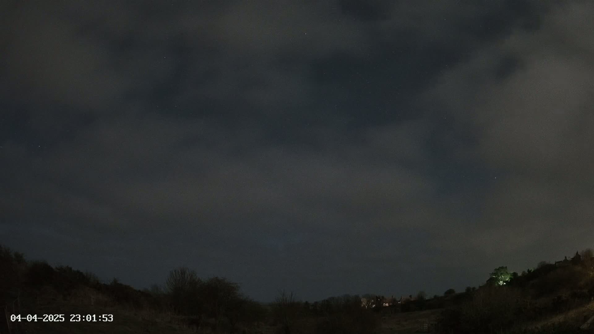 A nighttime landscape shows a dark, cloudy sky over a hill dotted with dark trees and a few faintly lit buildings.