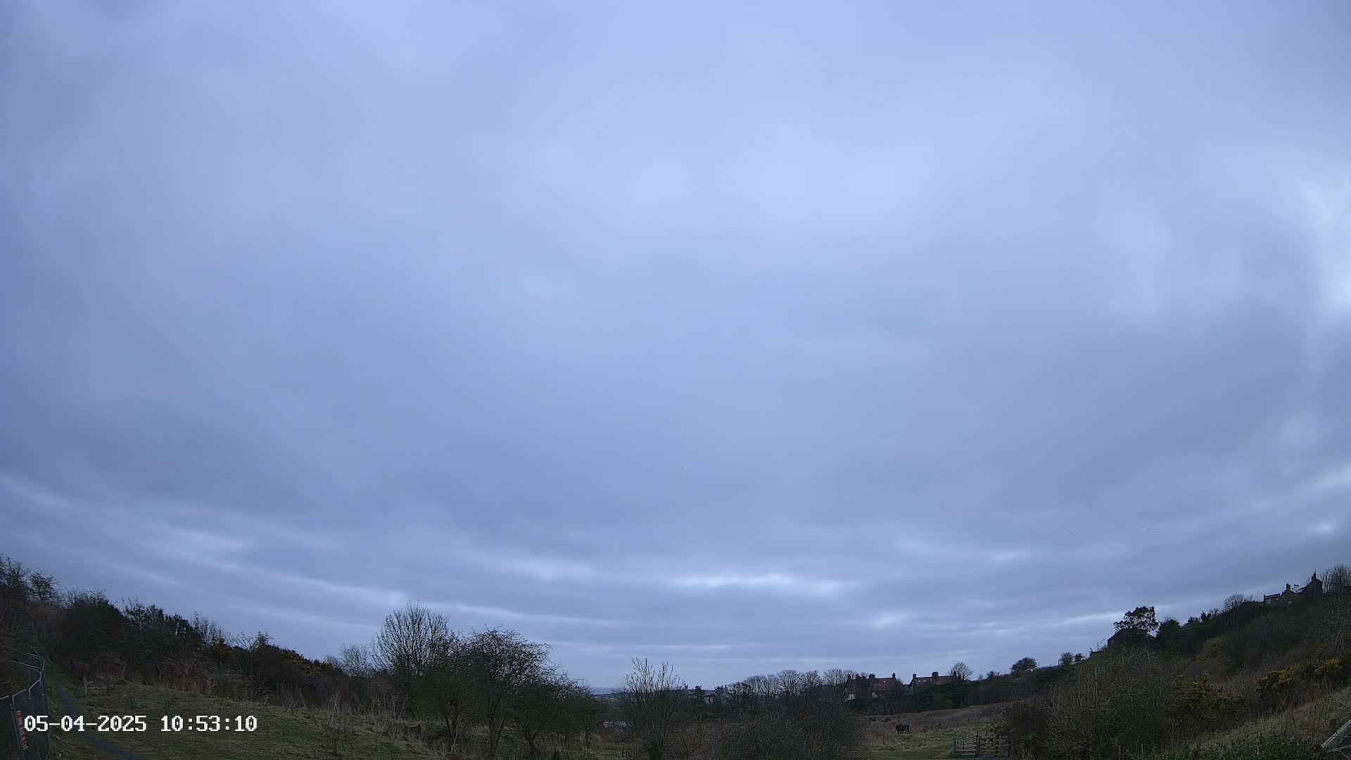 A mostly cloudy, pale blue sky overlooks a sparsely vegetated landscape with a few houses visible in the distance.