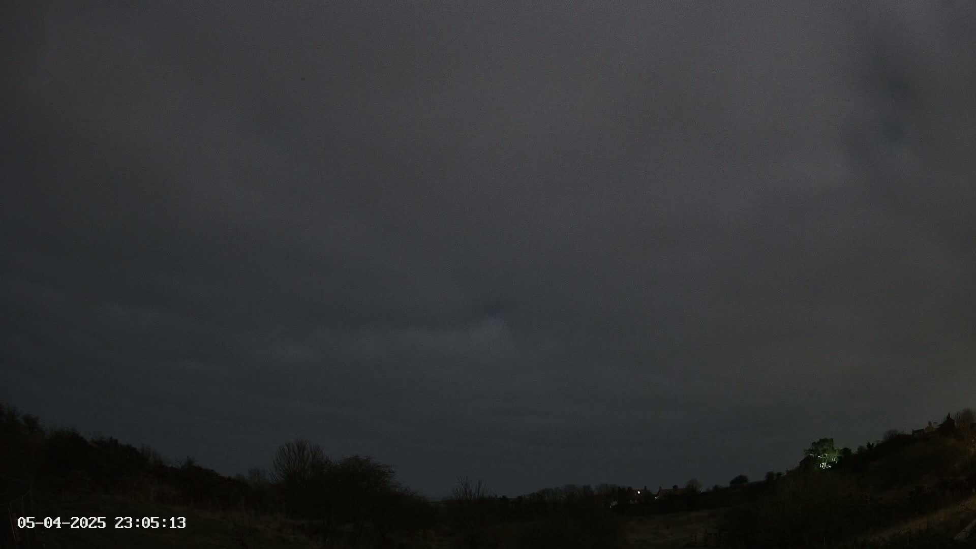 A dark, overcast night sky hangs over a silhouetted landscape of trees and a distant hilltop with a few lights.