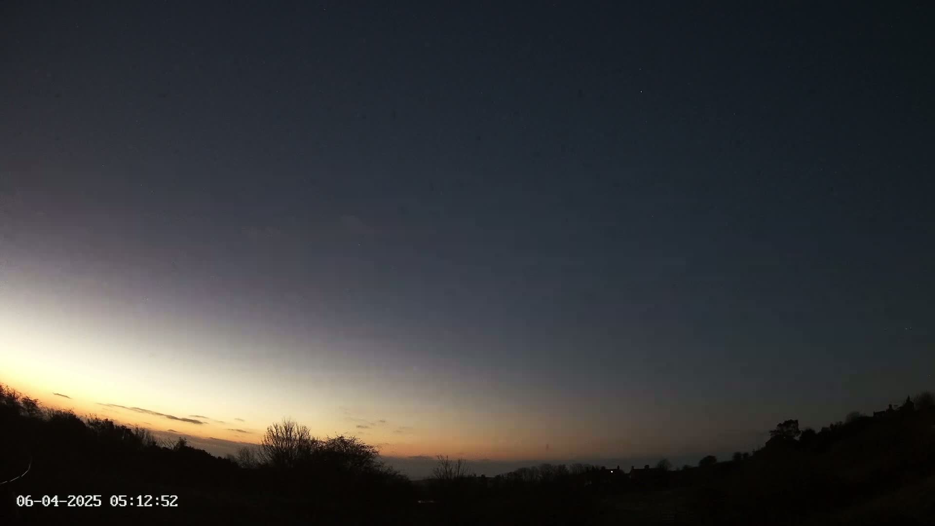 A dark sky transitioning to a pale orange and yellow sunrise over a silhouetted landscape of trees and buildings.