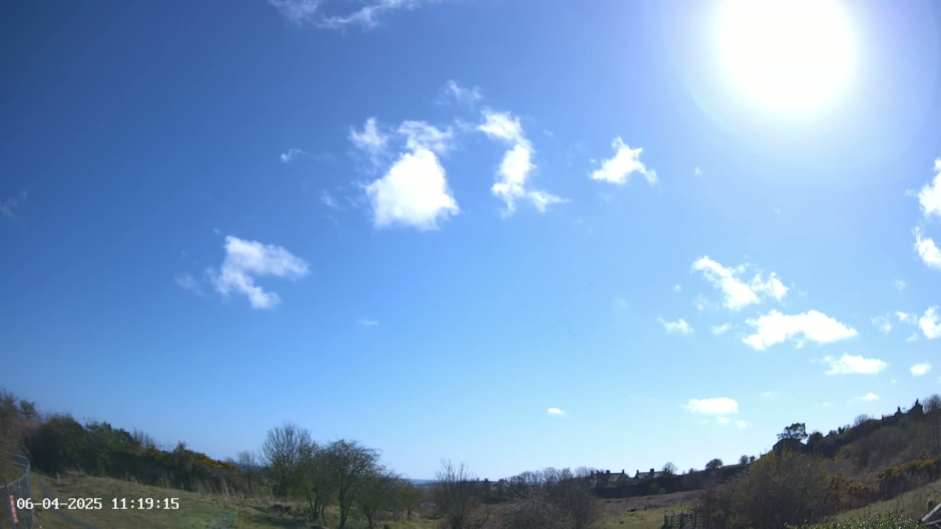 A sunny day with a mostly clear blue sky and scattered fluffy clouds over a landscape of low-lying vegetation and a few distant buildings.