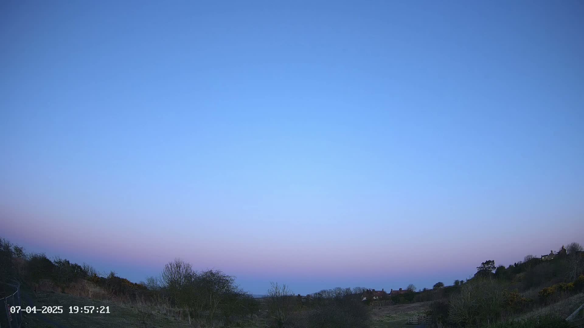 A clear, twilight sky displays soft purple and blue hues over a rural landscape with bare trees and distant buildings.
