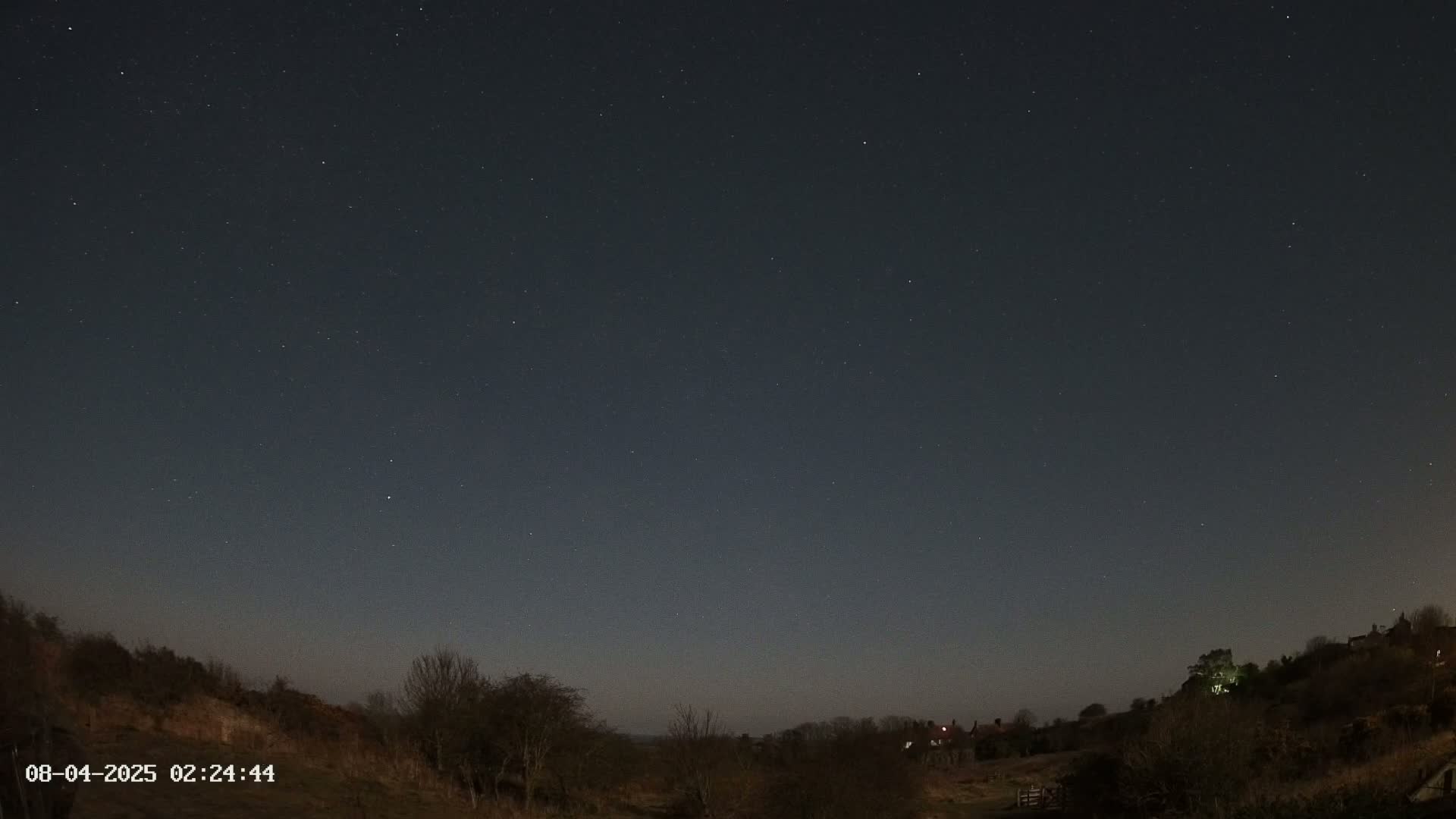 A mostly clear night sky filled with stars over a dark, hilly landscape with sparse, leafless trees.