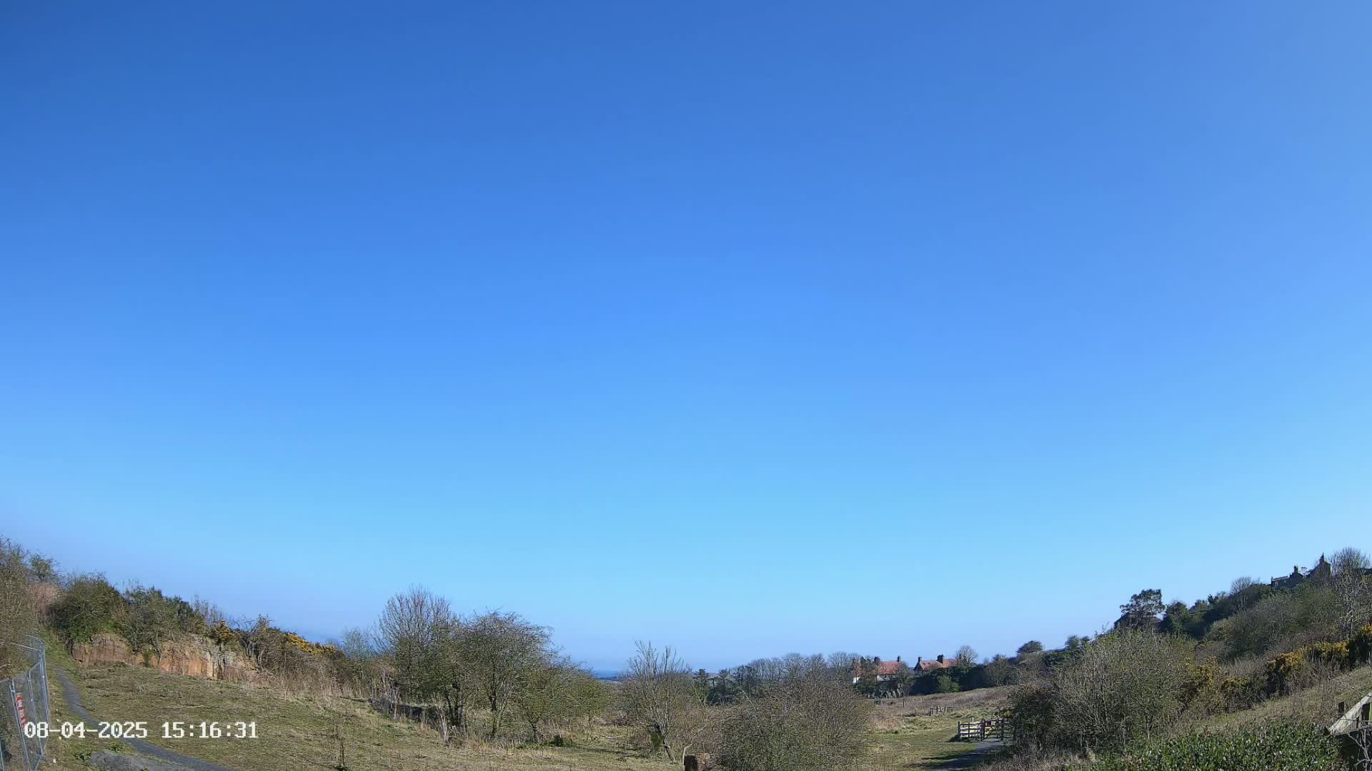 Under a clear blue sky, a grassy field gently slopes down towards a line of houses nestled among bare trees and shrubs.