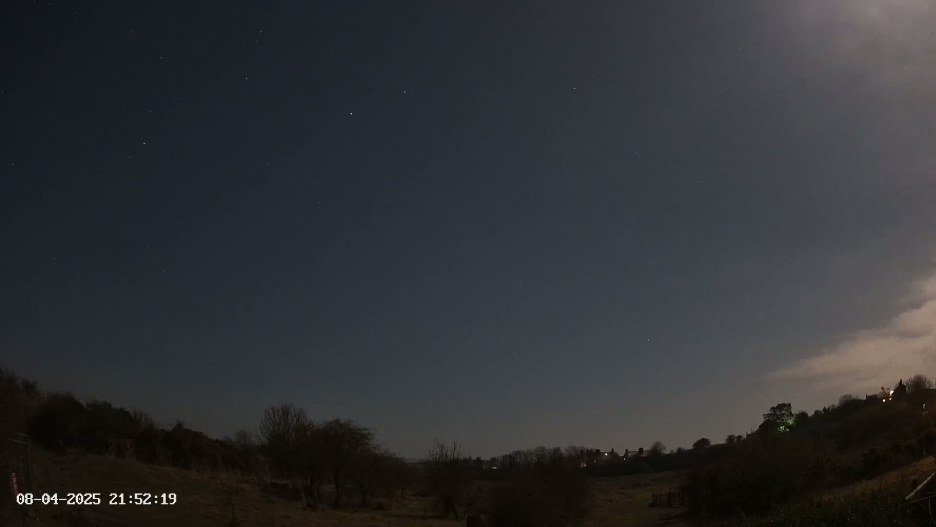 A night-time view of a dark, mostly clear sky with a few stars visible, overlooking a sparsely vegetated field and distant houses under a slightly cloudy moonlit sky.