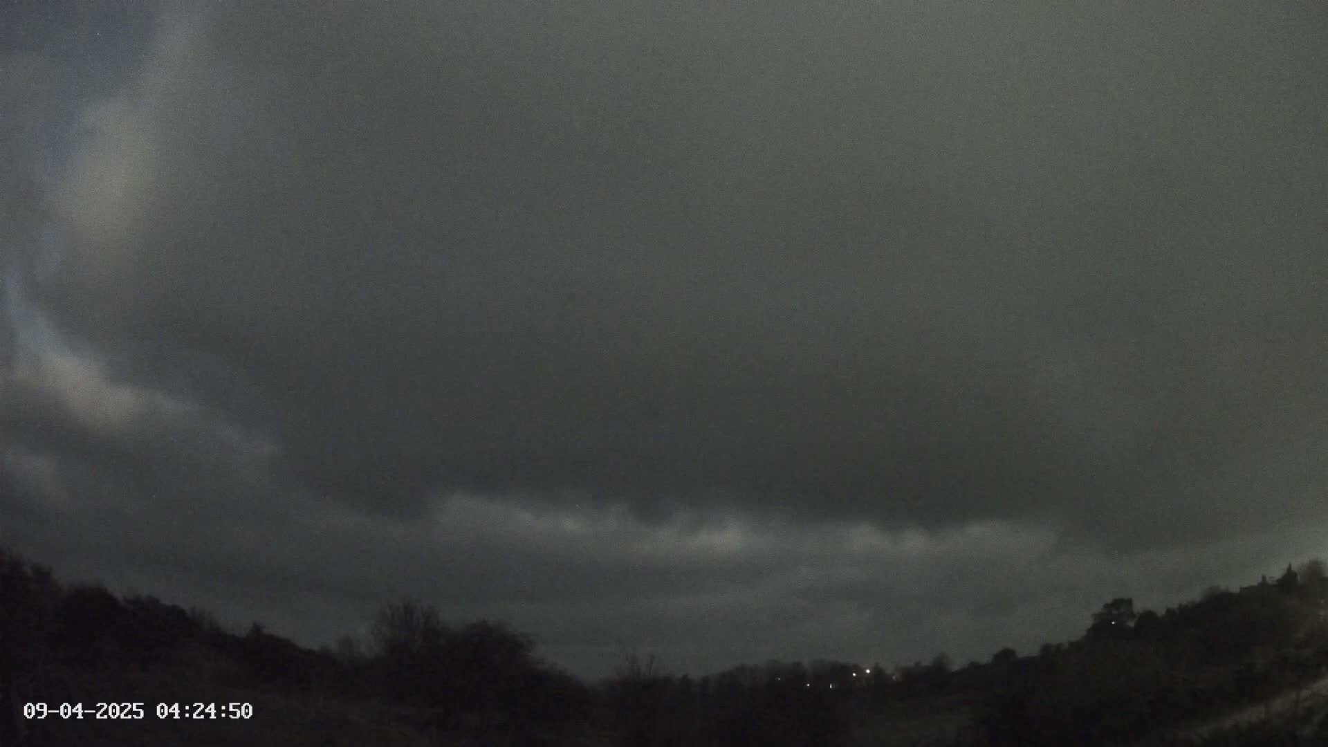 A dark, overcast sky hangs low over a silhouetted landscape of trees and hills at night.