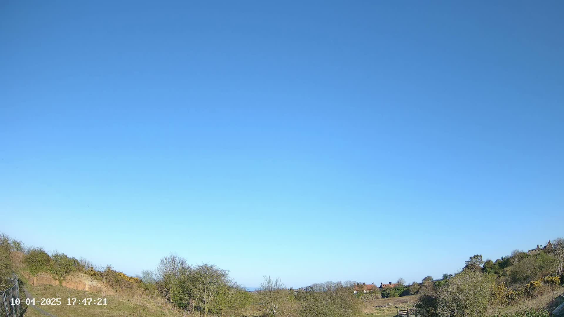 Under a clear blue sky, a hilly landscape with sparse trees and a few distant buildings is visible.