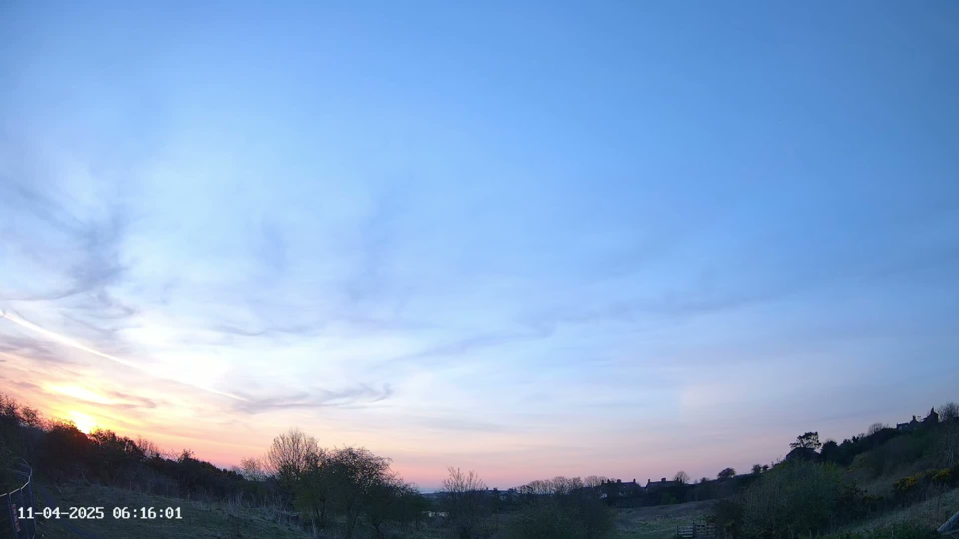 A sunset with a mostly clear, light-blue sky displays pastel pink and orange hues at the horizon over a hilly landscape with silhouetted trees and buildings.