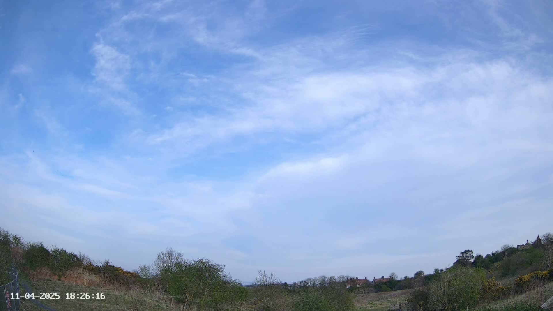 Under a mostly clear, light blue sky with thin, wispy clouds, a hilly landscape with sparse vegetation and a few distant houses is visible.