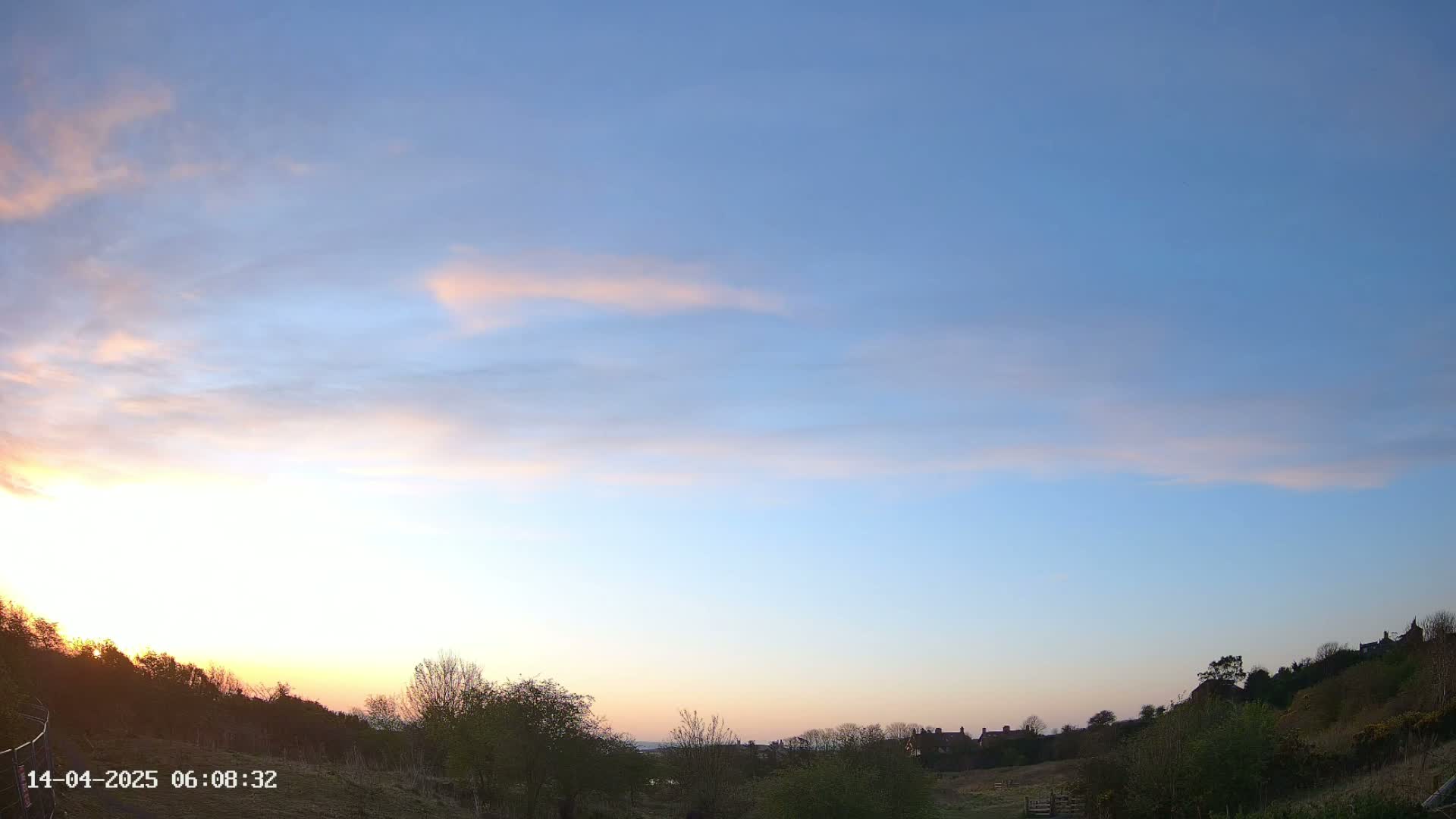 A sunrise with pale pink and orange clouds in a mostly clear blue sky over a landscape of trees and distant houses.