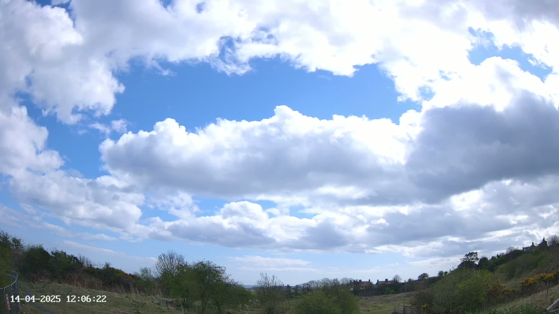 A mostly sunny sky filled with fluffy white cumulus clouds overlooks a landscape of green trees and shrubs, with a few buildings visible in the distance.
