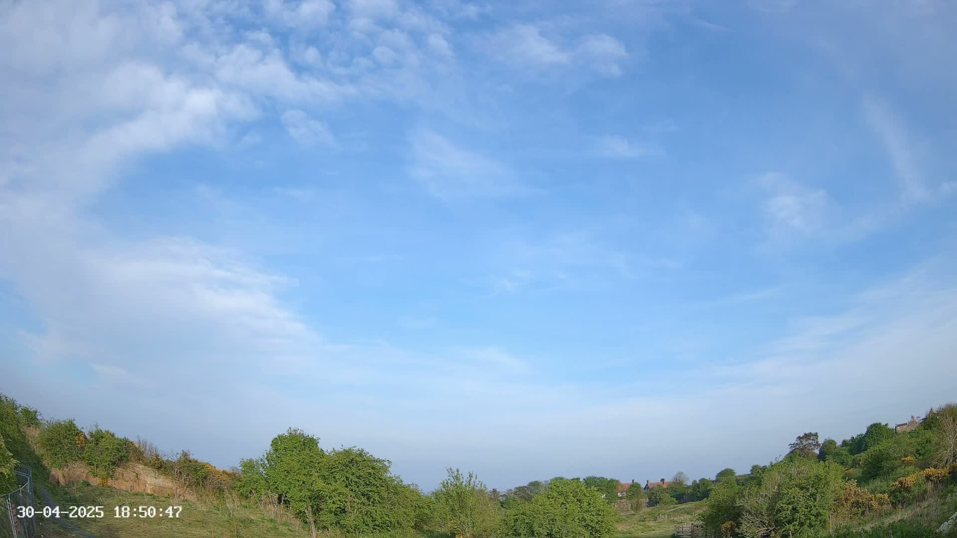 A mostly clear blue sky with scattered clouds overlooks a gently sloping landscape of green vegetation and distant houses.