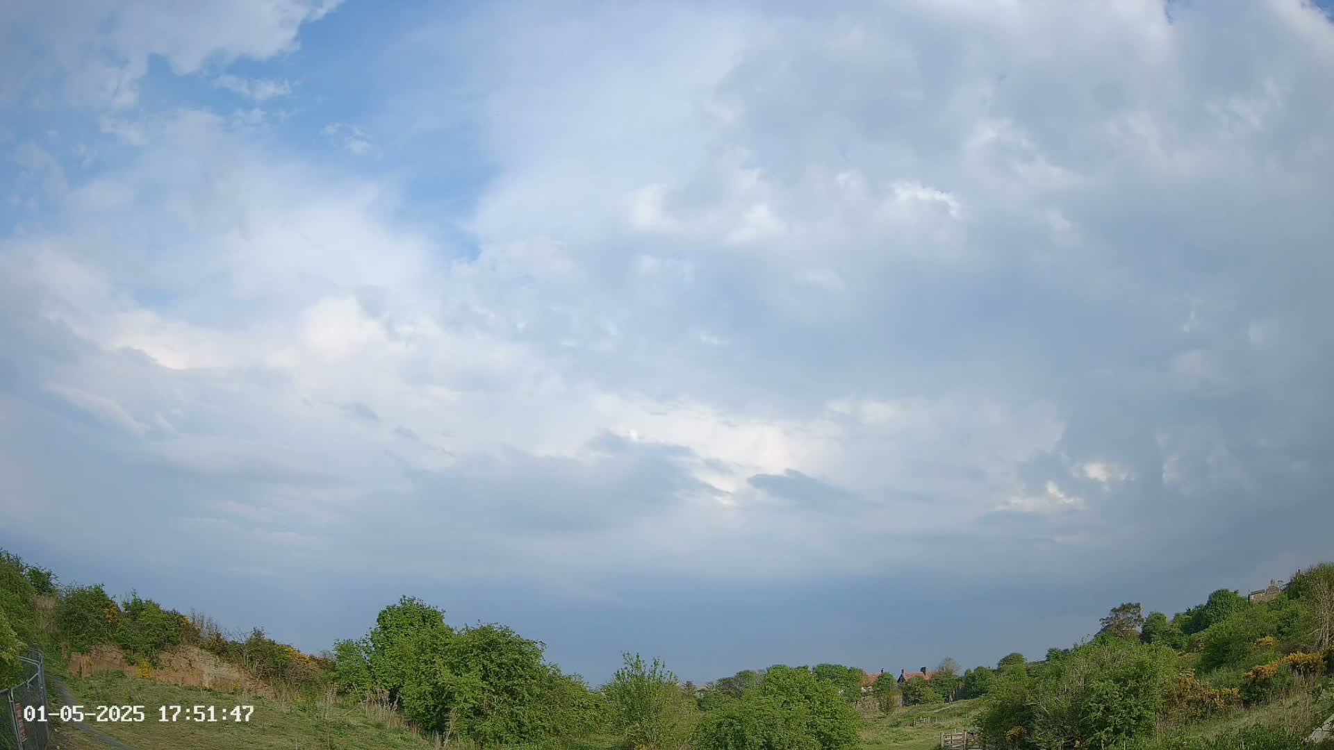 A mostly cloudy sky hangs over a gently sloping landscape of green trees and shrubs.
