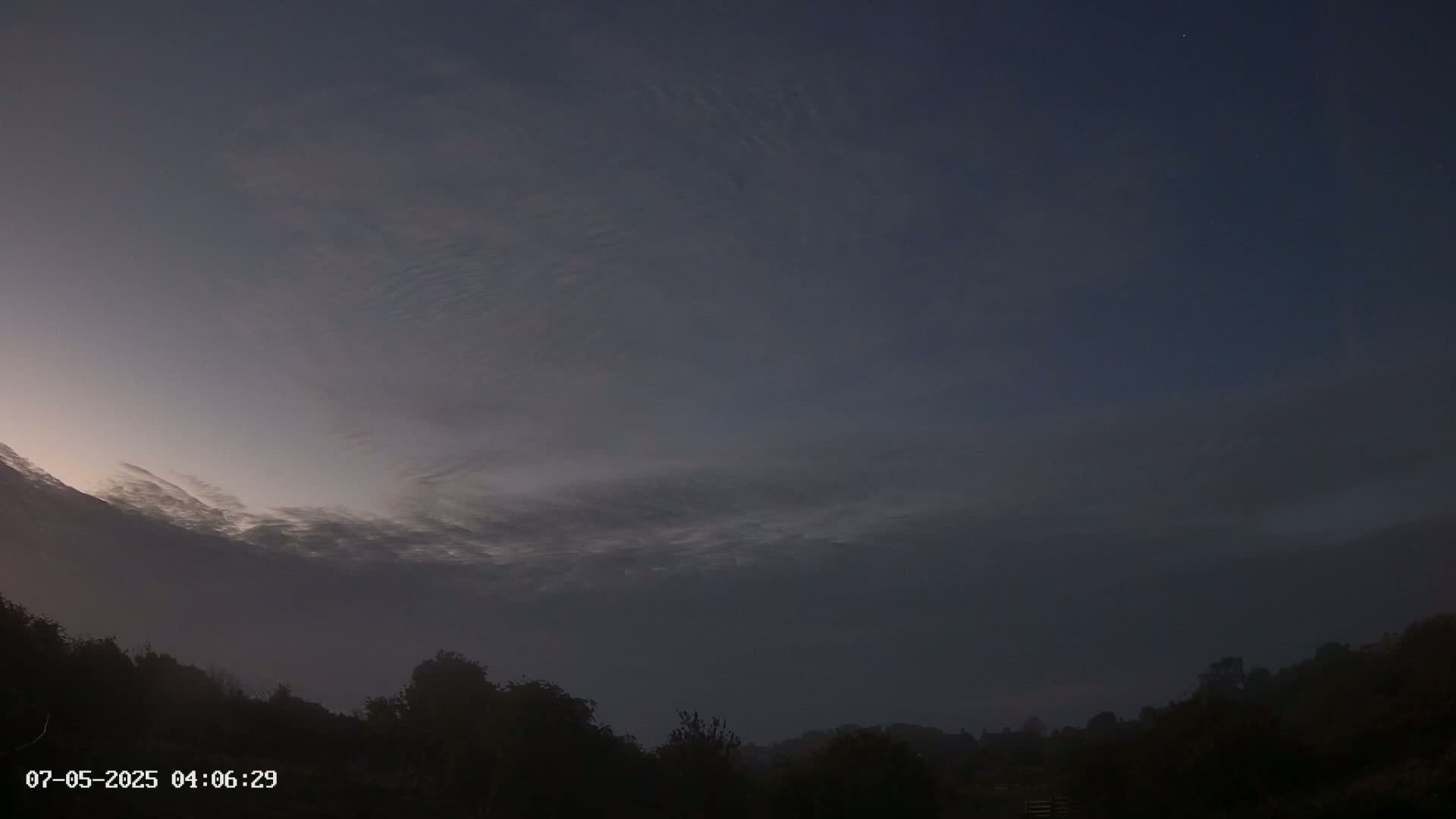 A foggy, pre-dawn sky shows thin, wispy clouds above a dark, silhouetted treeline.