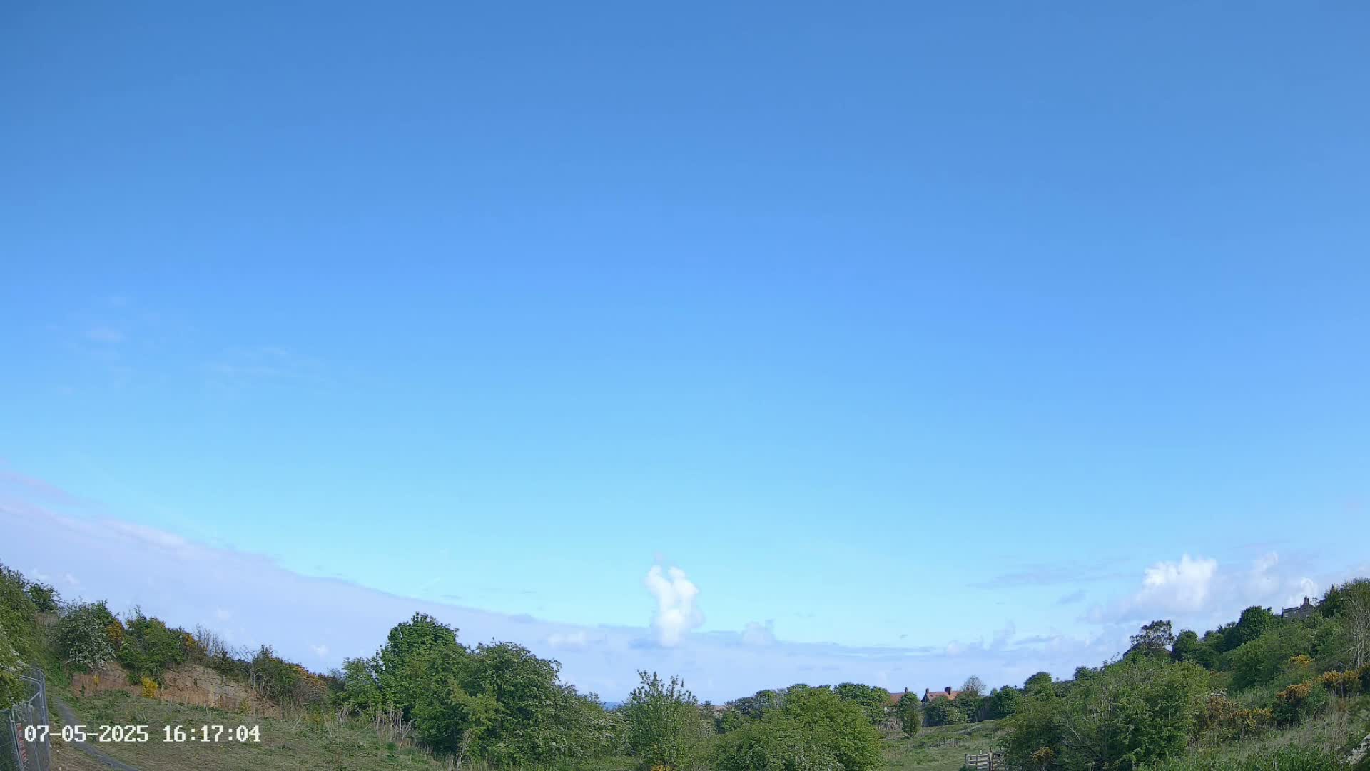 A mostly clear blue sky over a gently rolling landscape of green vegetation, with a few small, scattered clouds.