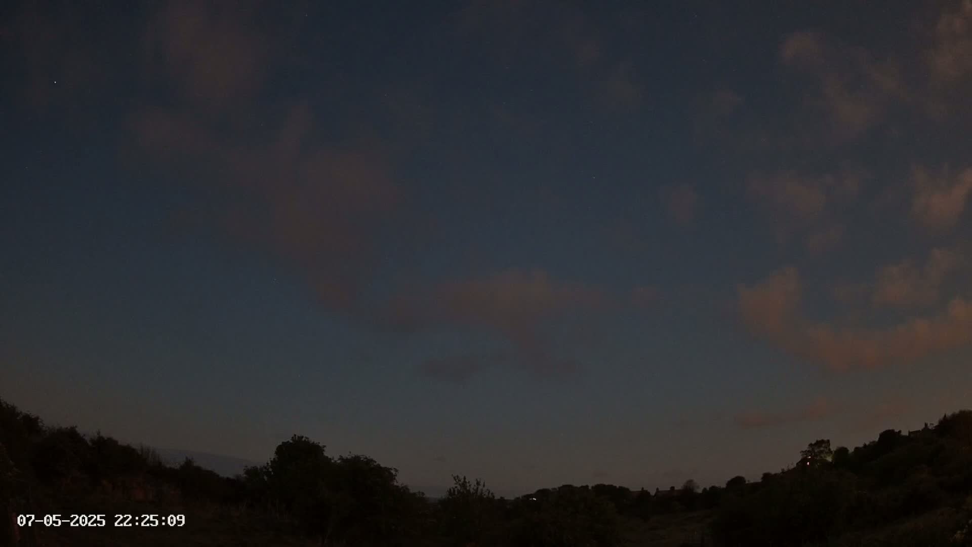 A nighttime view of a dark, mostly cloudy sky above a silhouetted landscape of trees and hills.