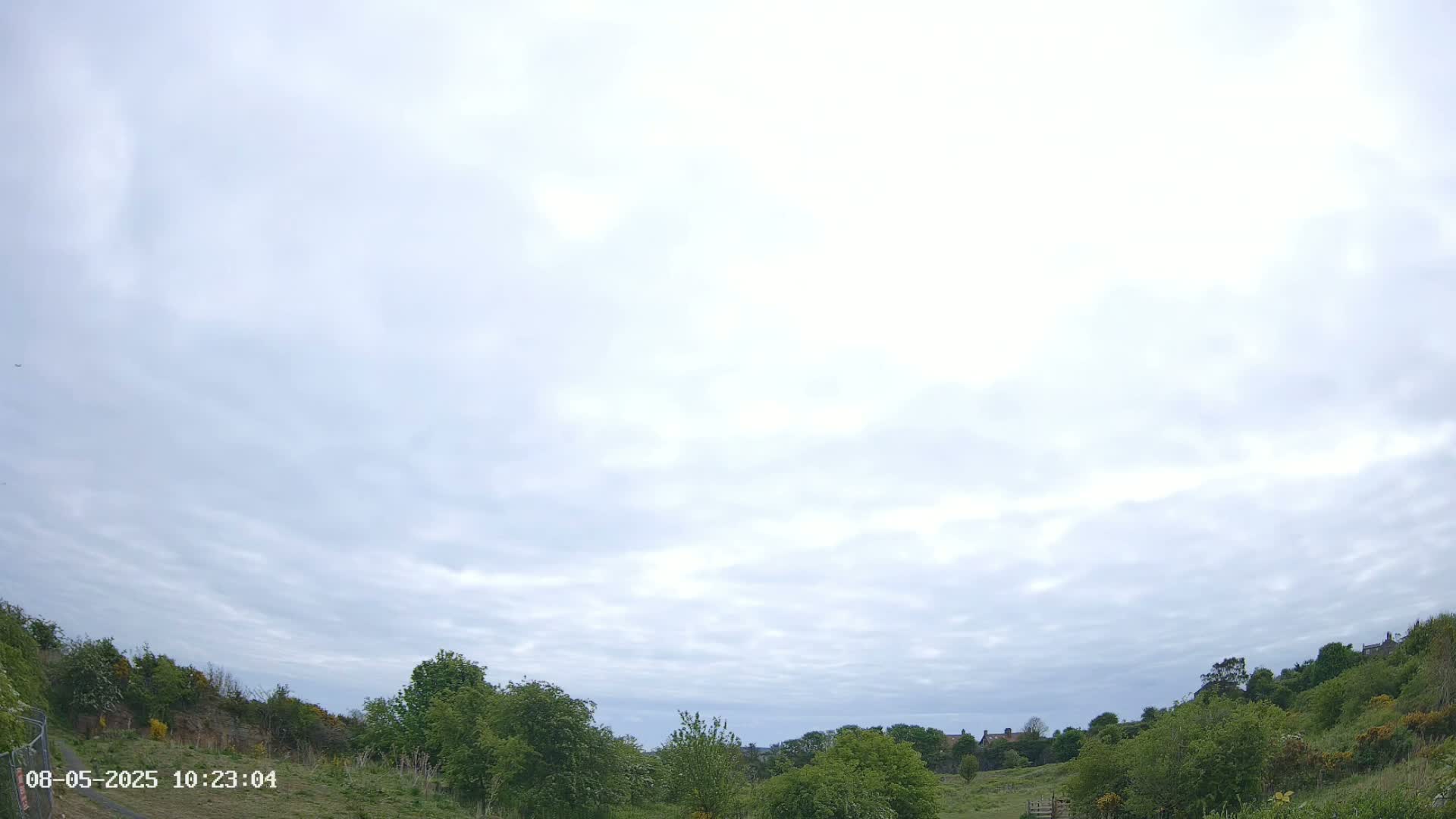Under a mostly cloudy sky, green hills covered with lush vegetation and trees stretch into the distance.