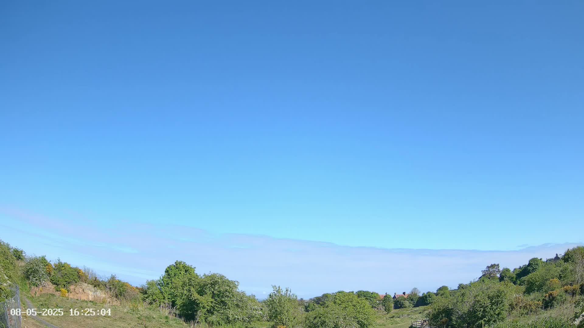 A mostly clear blue sky with a thin layer of low-lying clouds above a hilly landscape of green trees and shrubs.