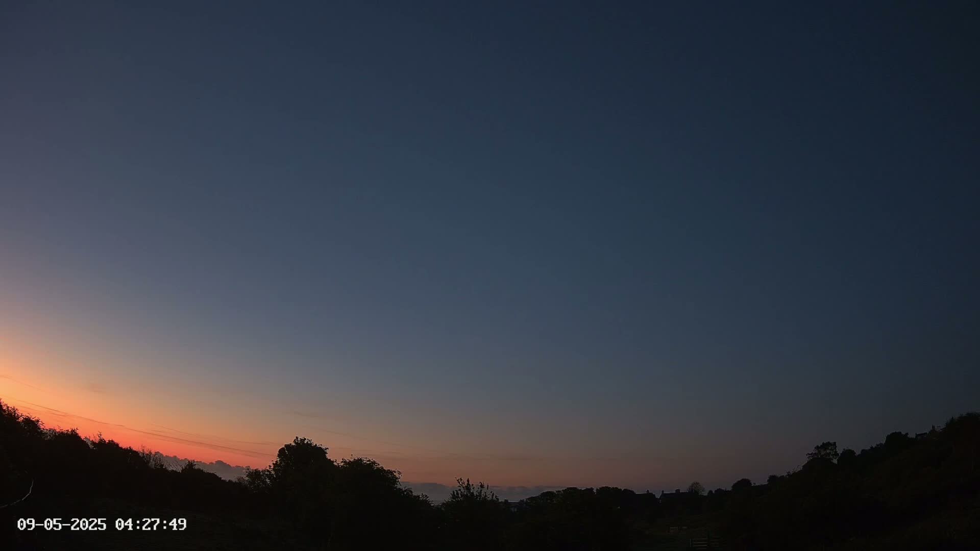 A sunrise with a mostly clear sky illuminates the silhouettes of trees and distant land.