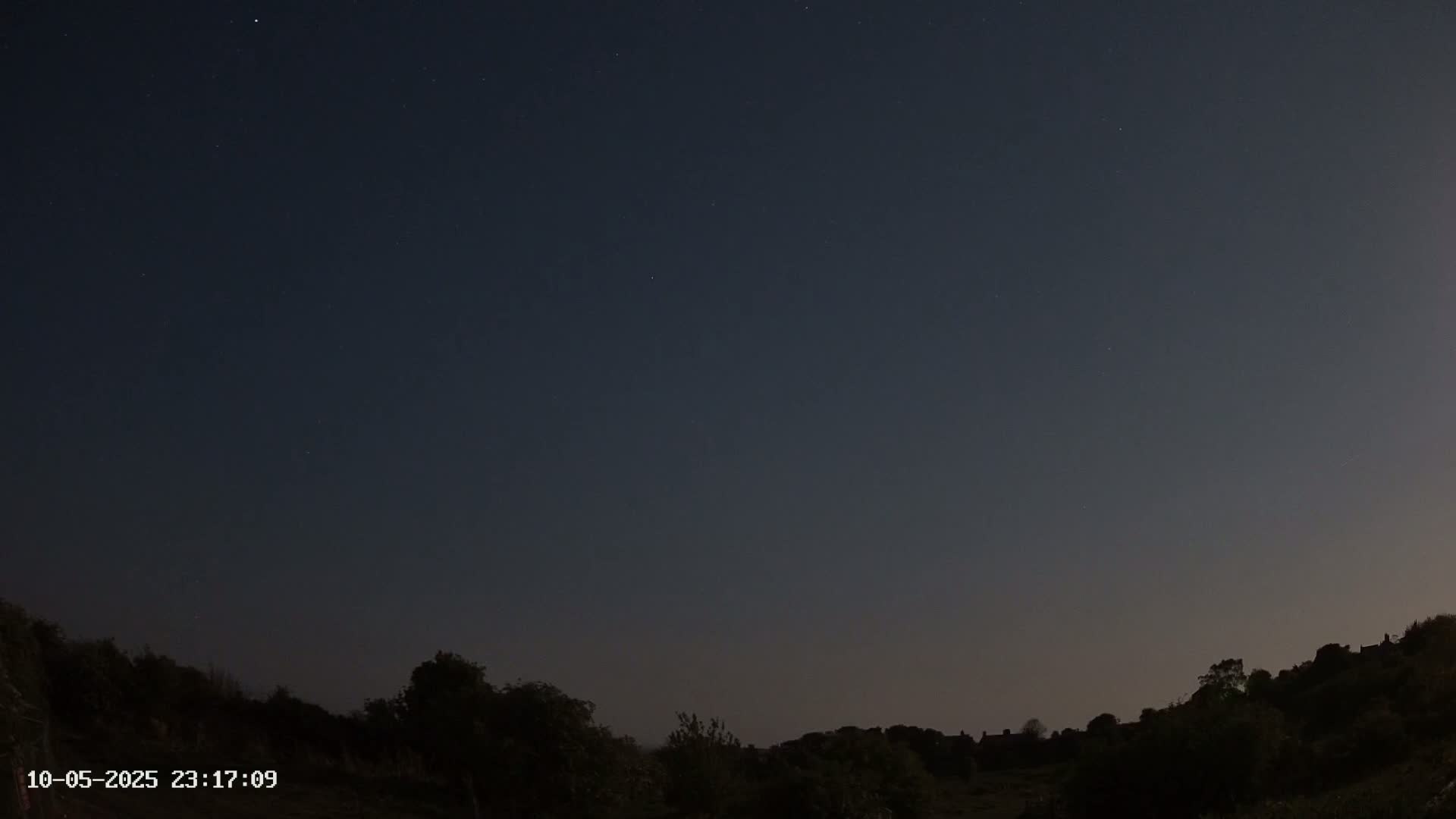 A night sky with a few visible stars over a dark, silhouetted landscape of trees and hills.