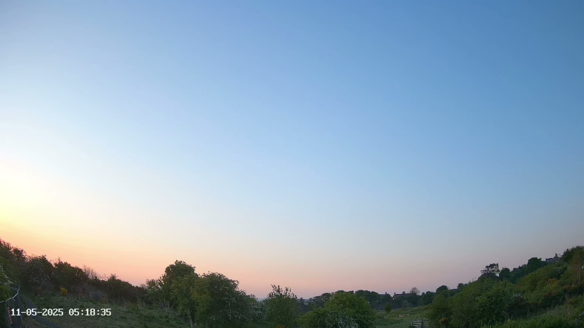 A pale pink and blue sunrise illuminates a landscape of green hills and trees.