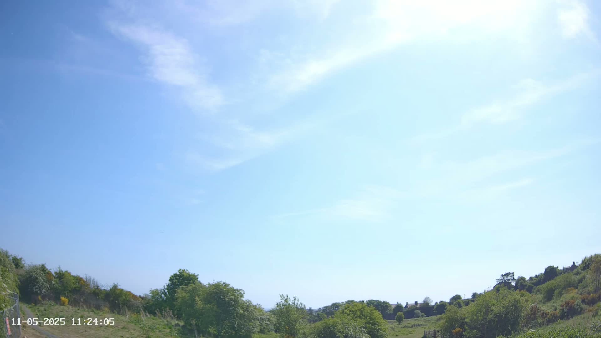 A mostly sunny sky with a few wispy clouds overlooks a hilly landscape of green vegetation.