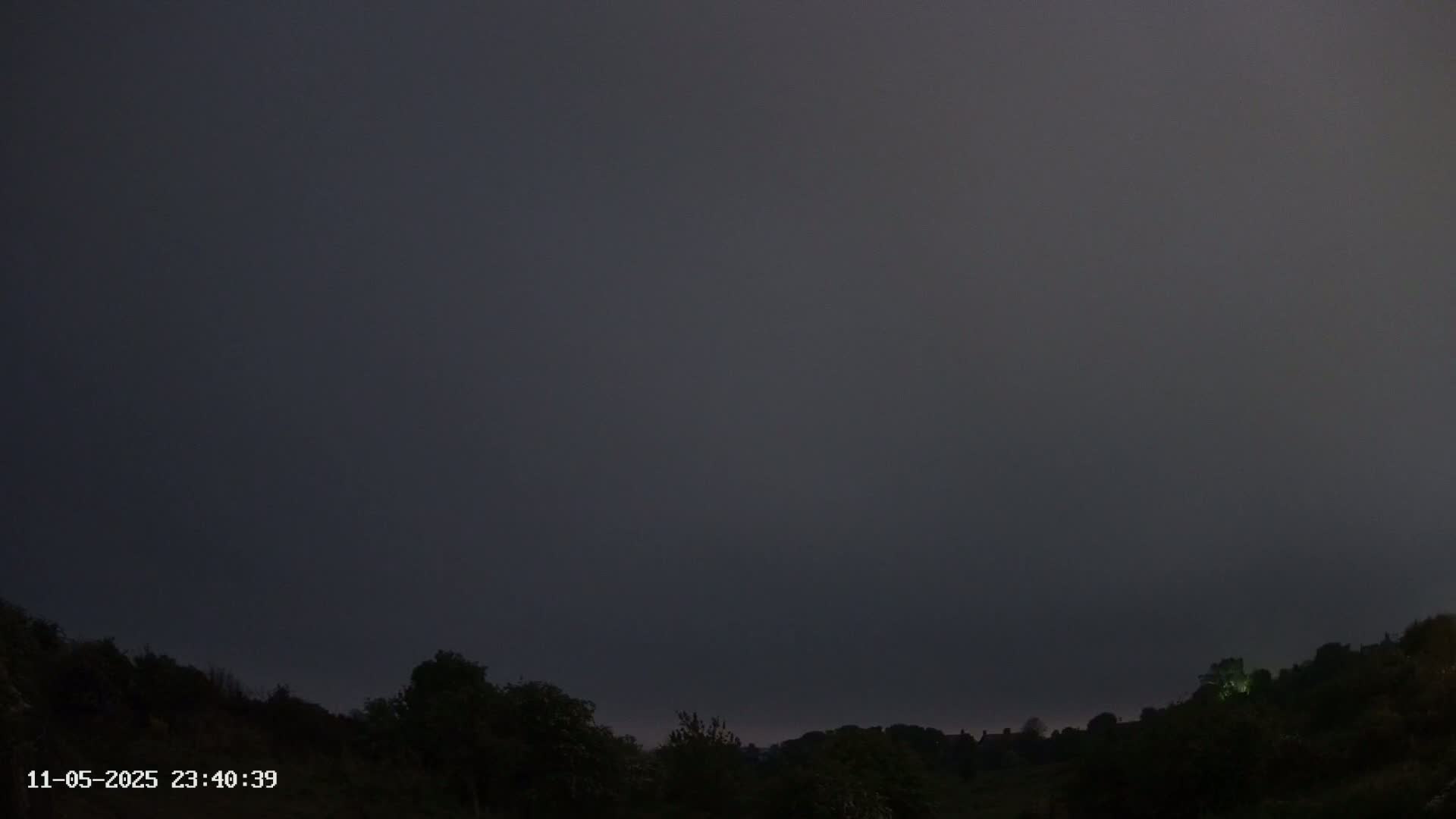 A dark, overcast sky hangs over a silhouetted line of trees and hills at night.