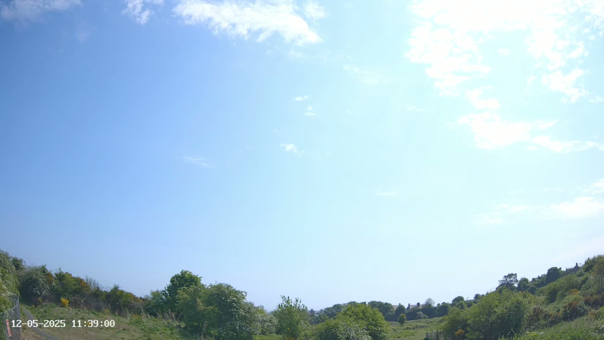 Under a mostly sunny sky with a few scattered clouds, green hills and vegetation are visible, with some buildings in the distance.