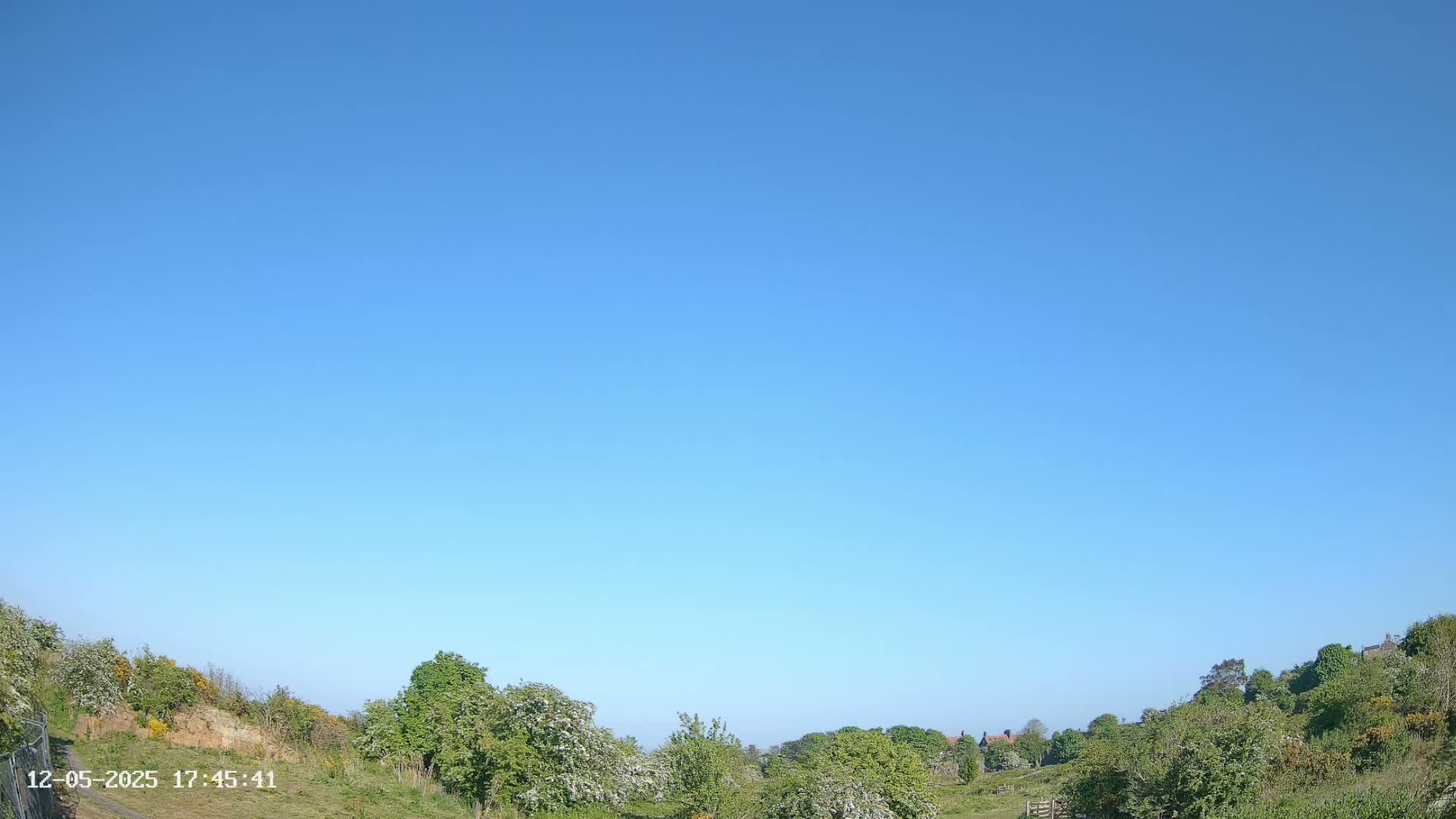 A clear blue sky overlooks a gently sloping landscape of green vegetation and a few distant buildings.