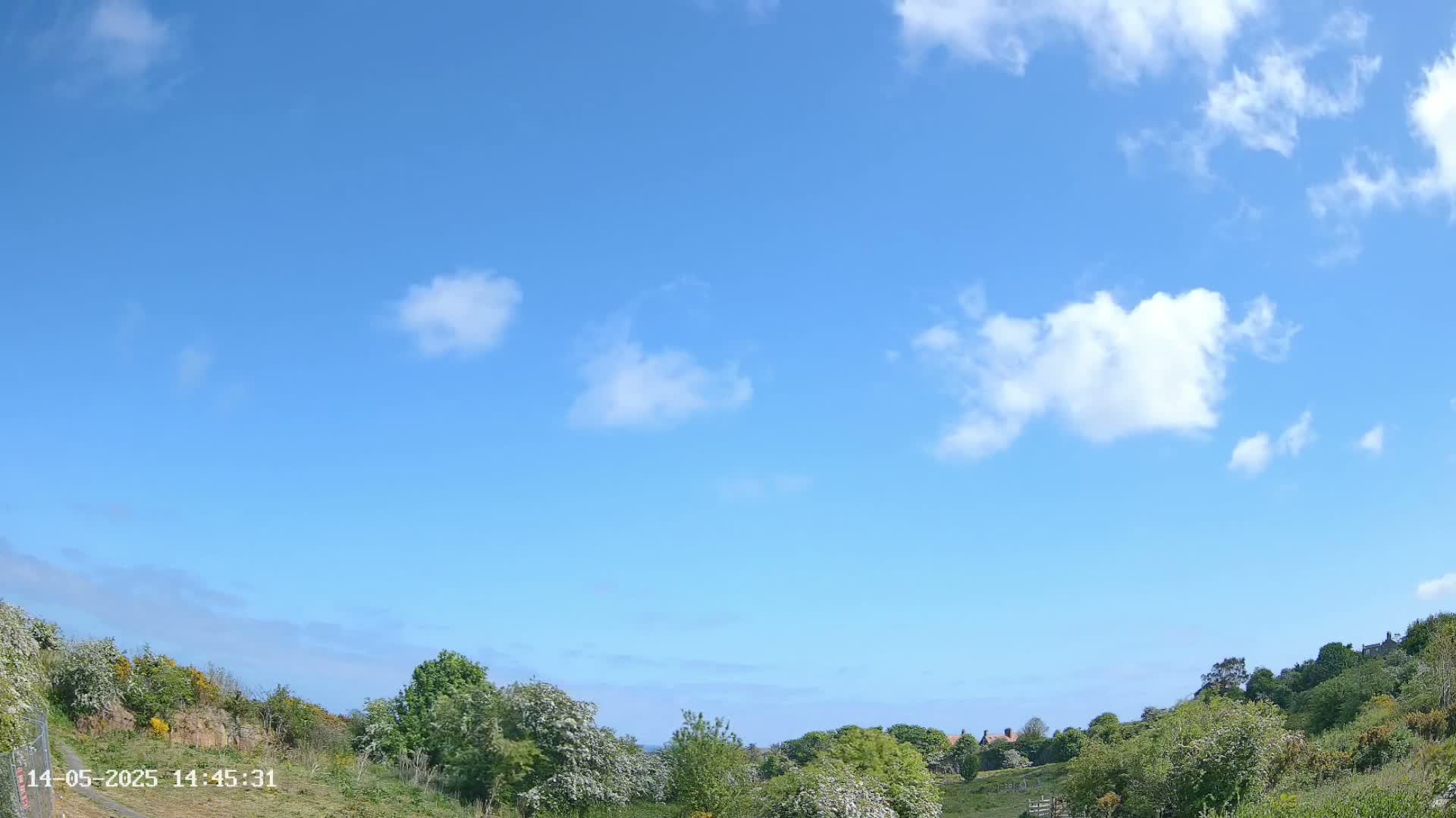 A mostly sunny day with scattered white clouds over a hilly landscape with green vegetation.