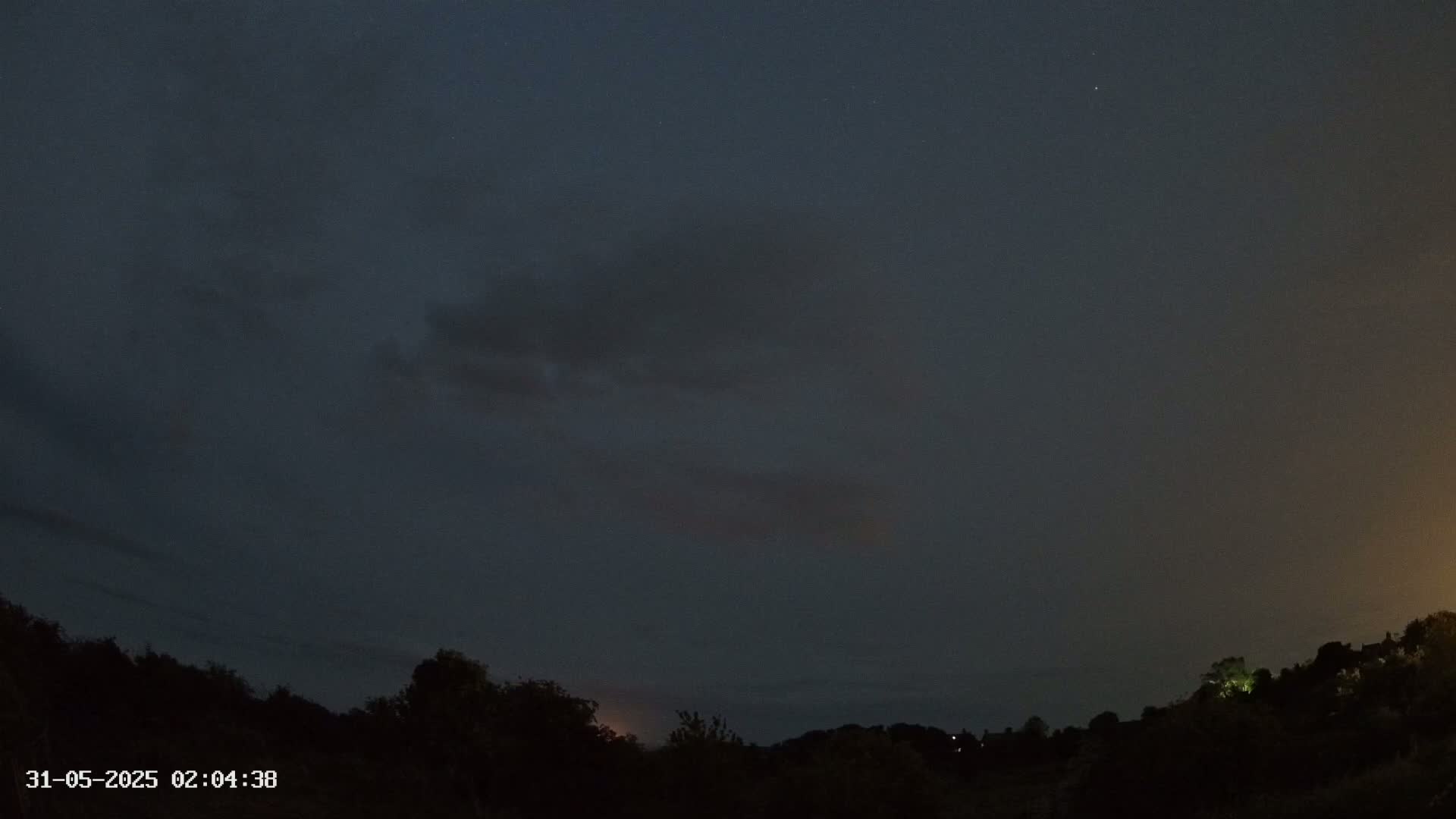 A dark, mostly cloudy night sky over a silhouetted landscape of trees and hills.