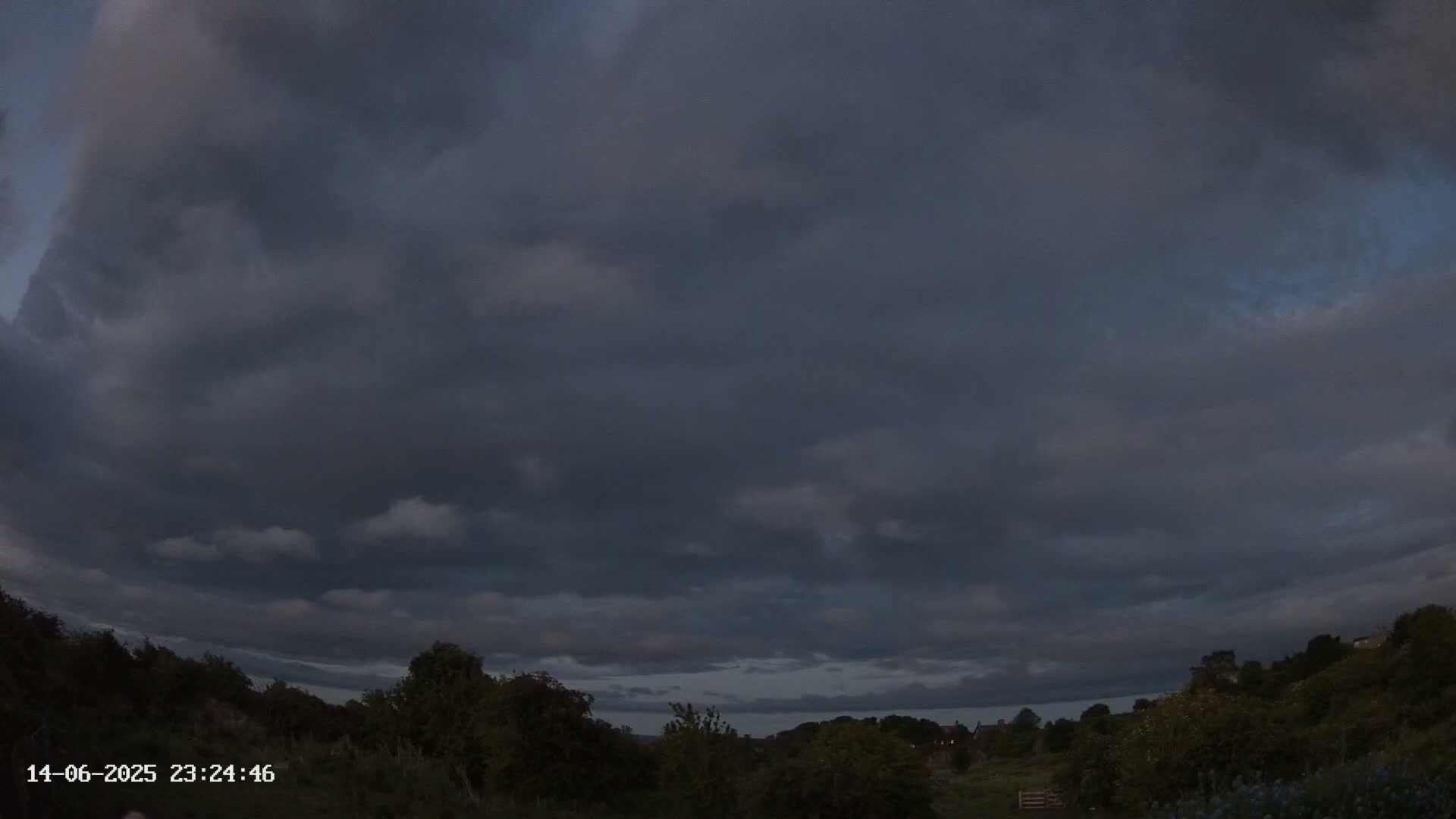 A mostly cloudy sky hangs over a green, hilly landscape with some shrubs and trees.