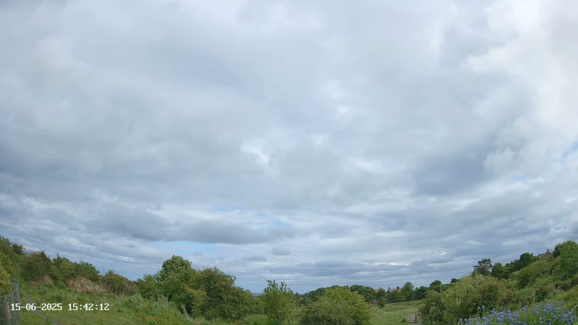 A mostly cloudy sky hangs over a gently sloping landscape of green vegetation, with a few distant buildings visible in the background.