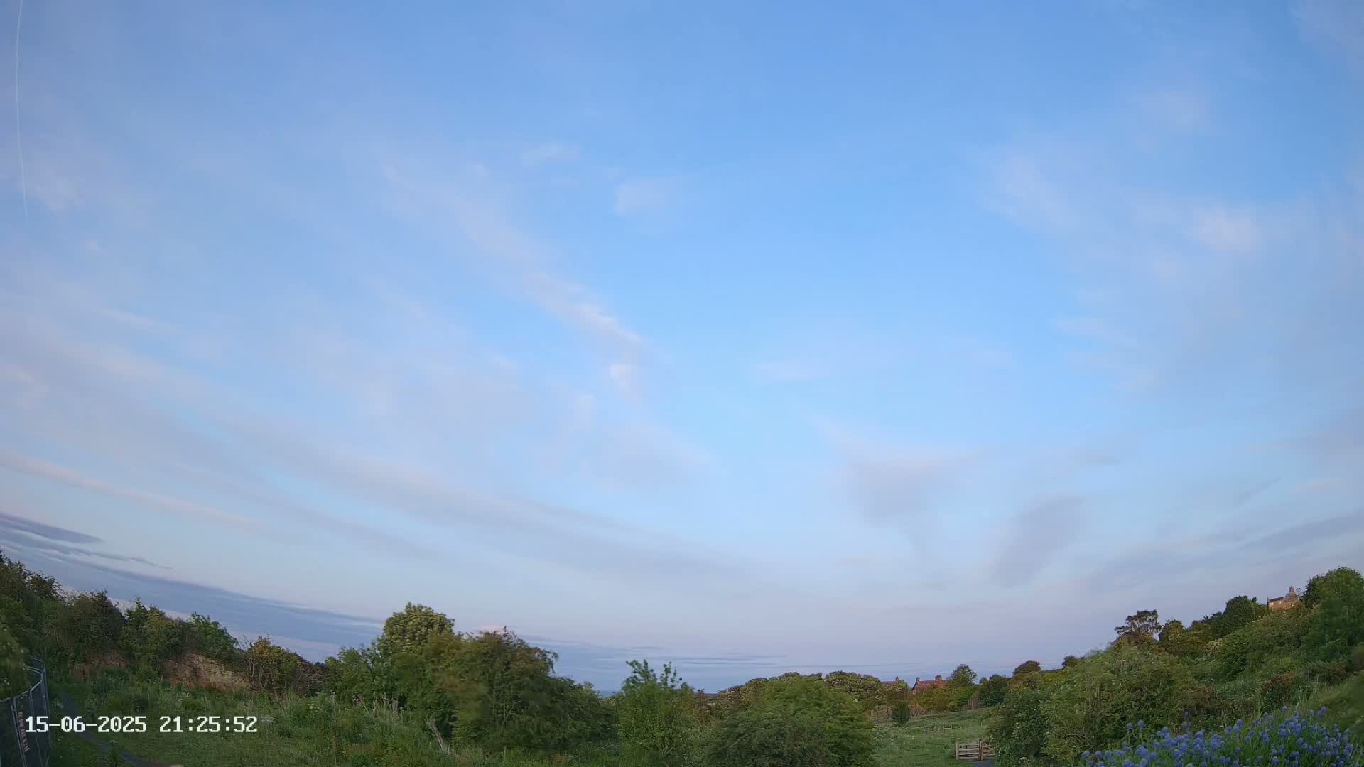 A mostly cloudy sky hangs over a gently sloping landscape of green vegetation, with a few distant buildings visible in the background.