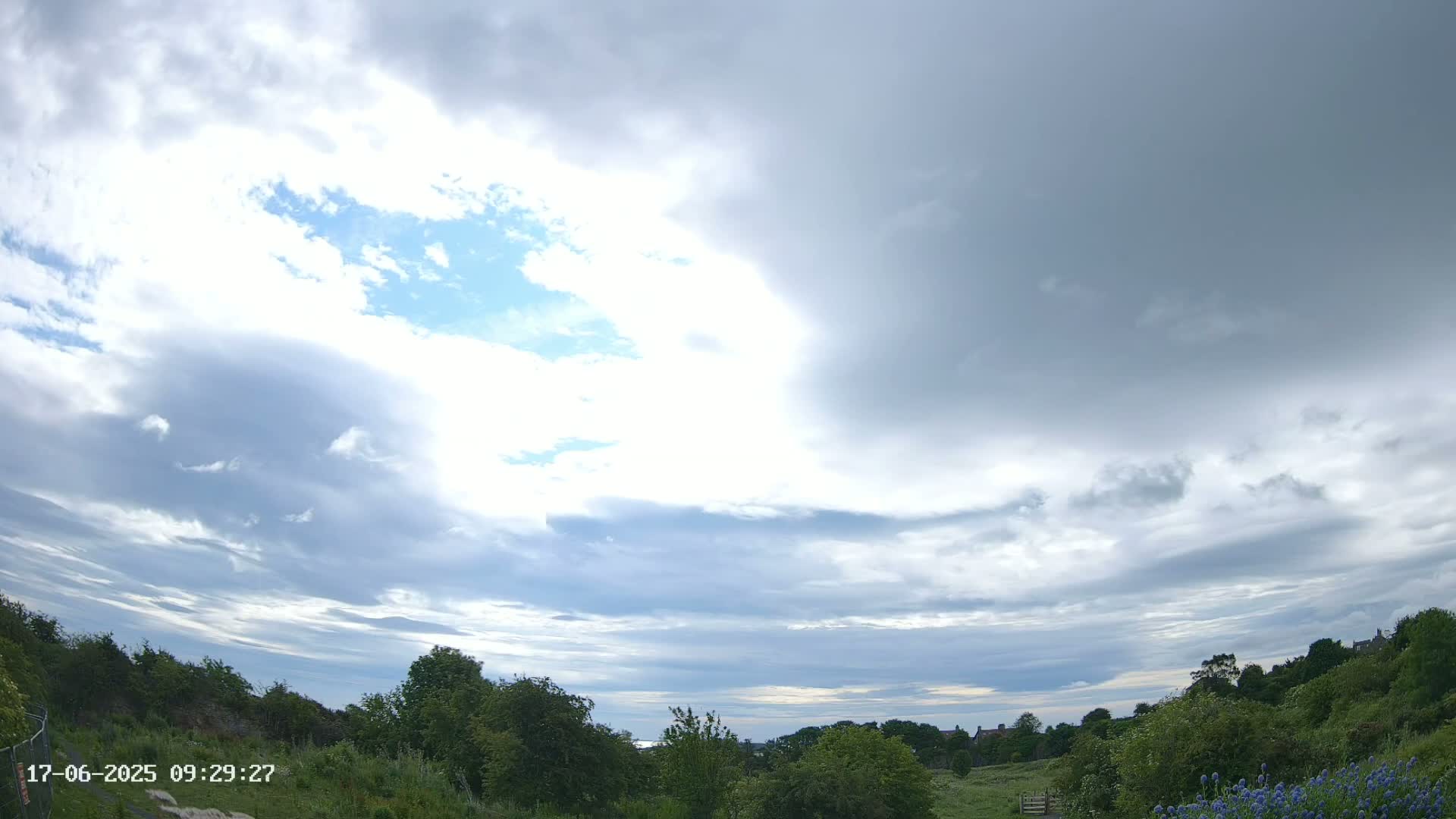 A partly cloudy sky over a green landscape with some distant houses.