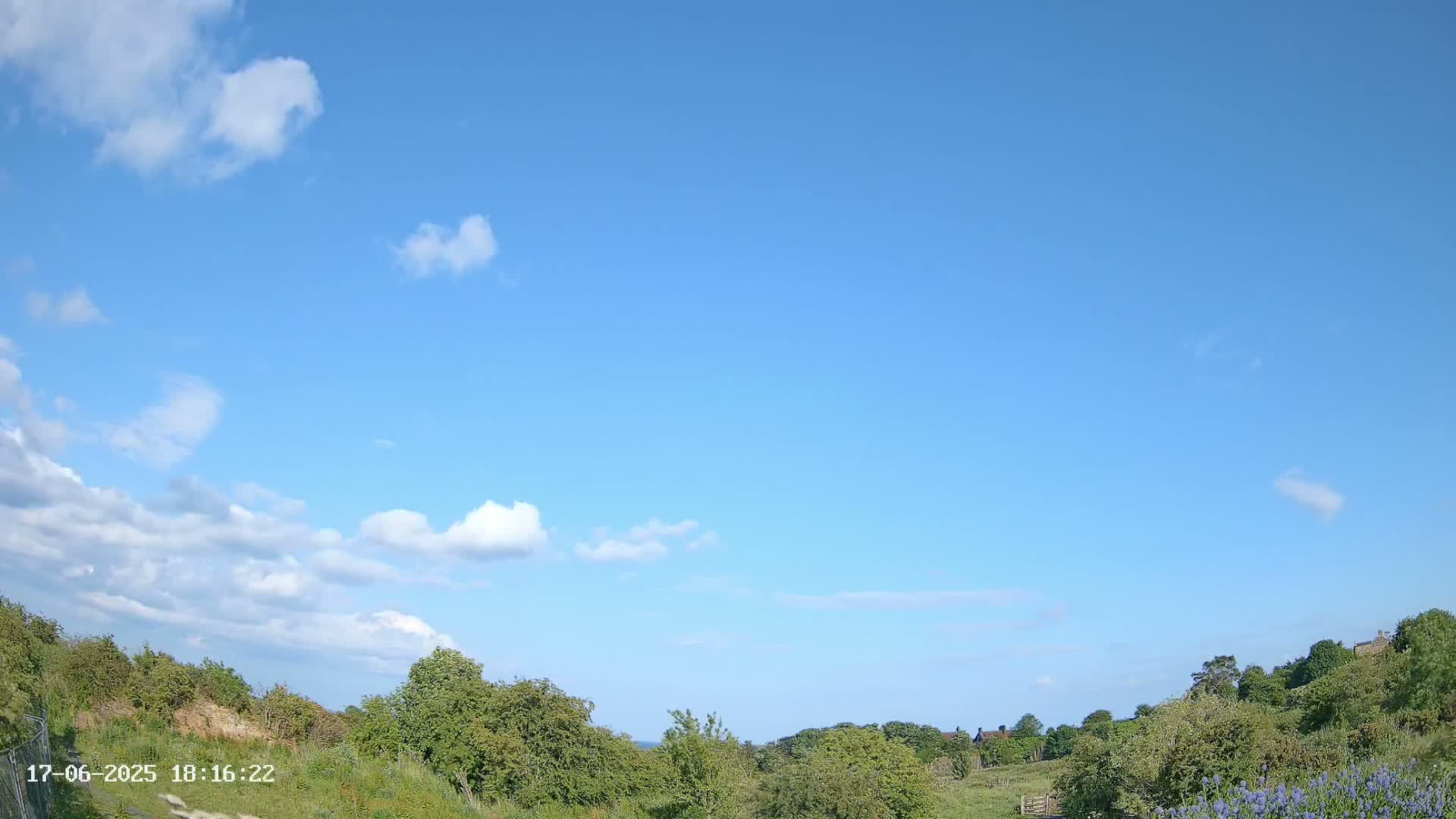 A mostly sunny sky with scattered cumulus clouds overlooks a gently rolling landscape of green vegetation and distant buildings.