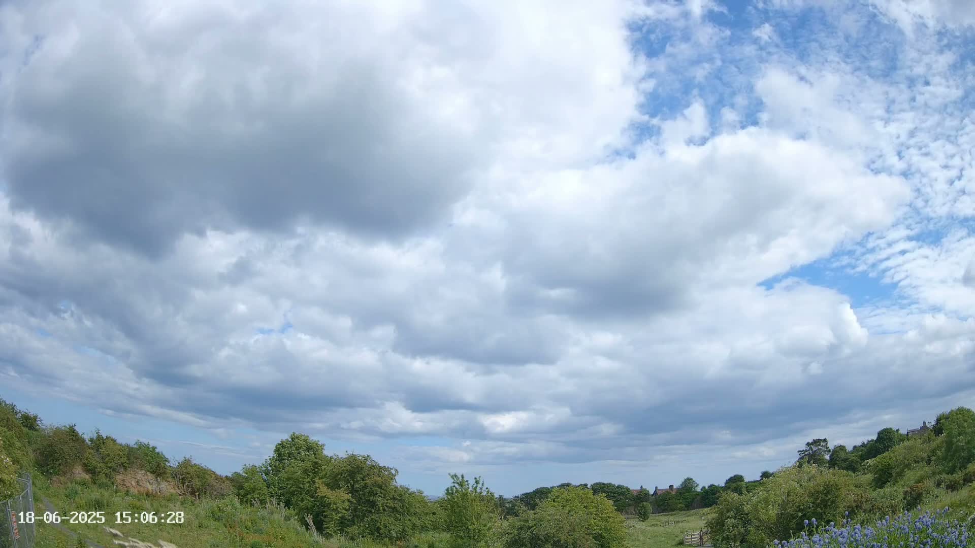 A partly cloudy sky over a verdant landscape with low-lying hills and some distant buildings.
