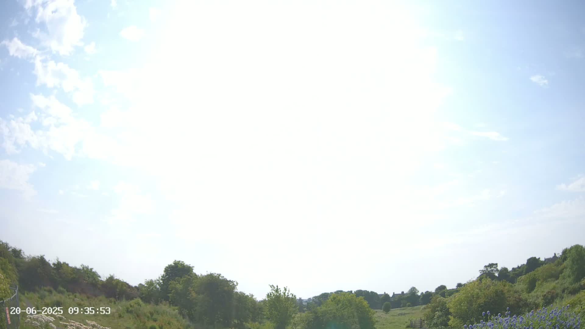 A sunny day with a mostly bright sky and scattered clouds over a green landscape of low hills and vegetation.