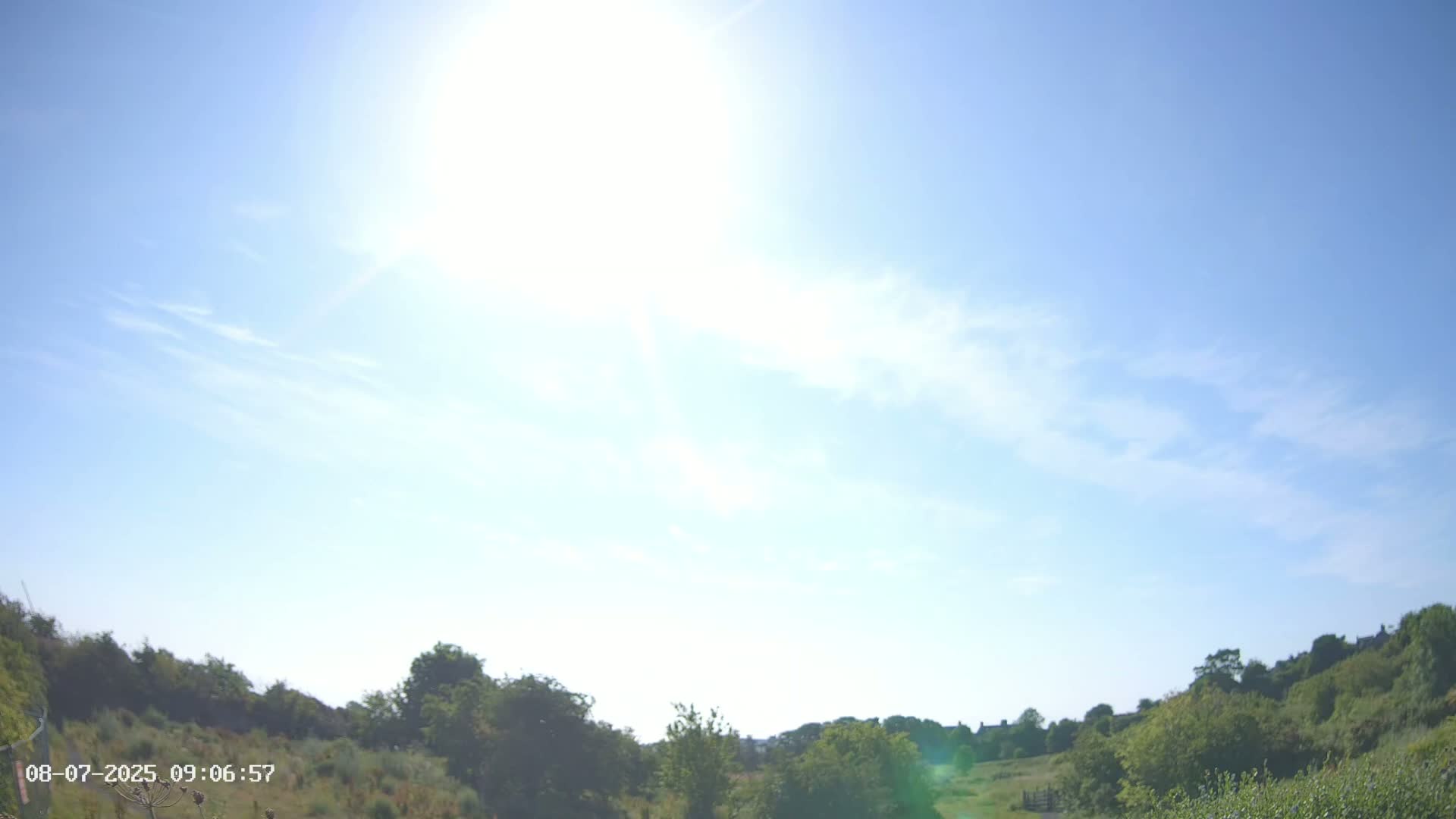 A sunny day with a mostly clear blue sky and some wispy clouds over a landscape of green shrubs and trees.