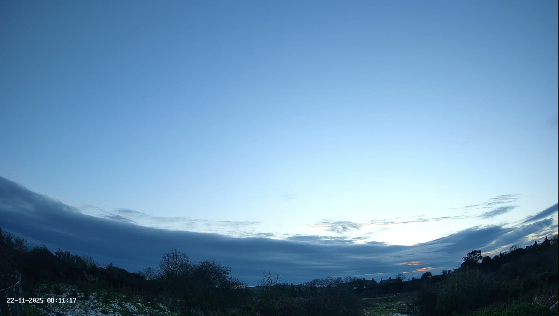A chilly, partially cloudy early morning landscape is depicted, showing a light dusting of snow on the ground, bare trees, and distant houses under a sky transitioning from deep blue to lighter tones with prominent dark cloud formations.