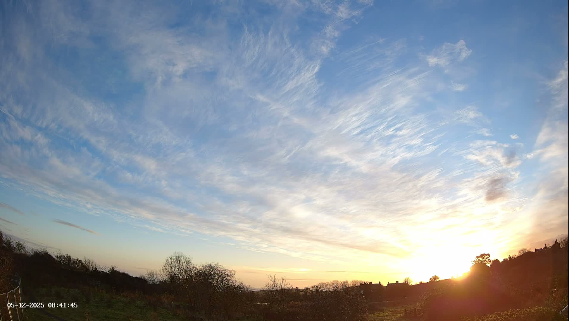 A brilliant sunrise illuminates a hilly landscape with bare trees and distant structures under a clear blue sky streaked with delicate wispy clouds, indicating fair weather.