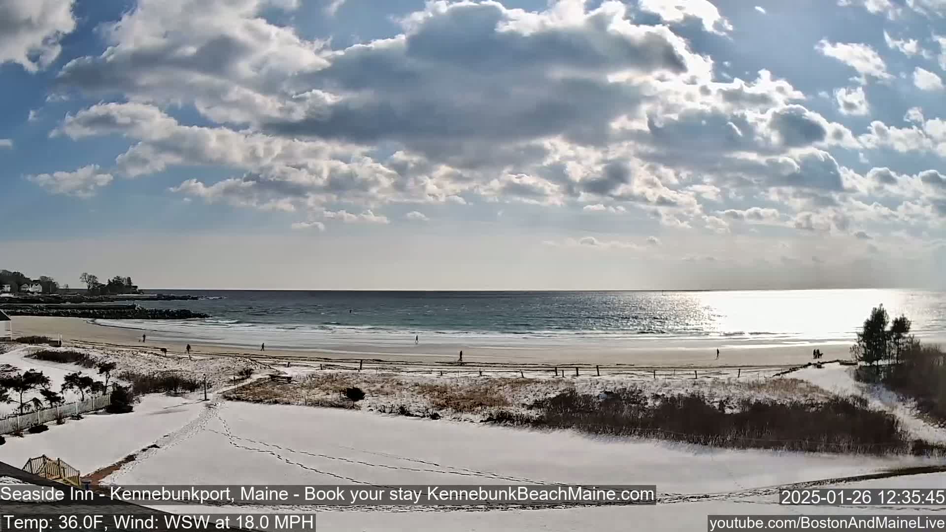 A partly sunny day shows a snowy beach with a few people walking along the shore and ocean waves, under a partly cloudy sky.